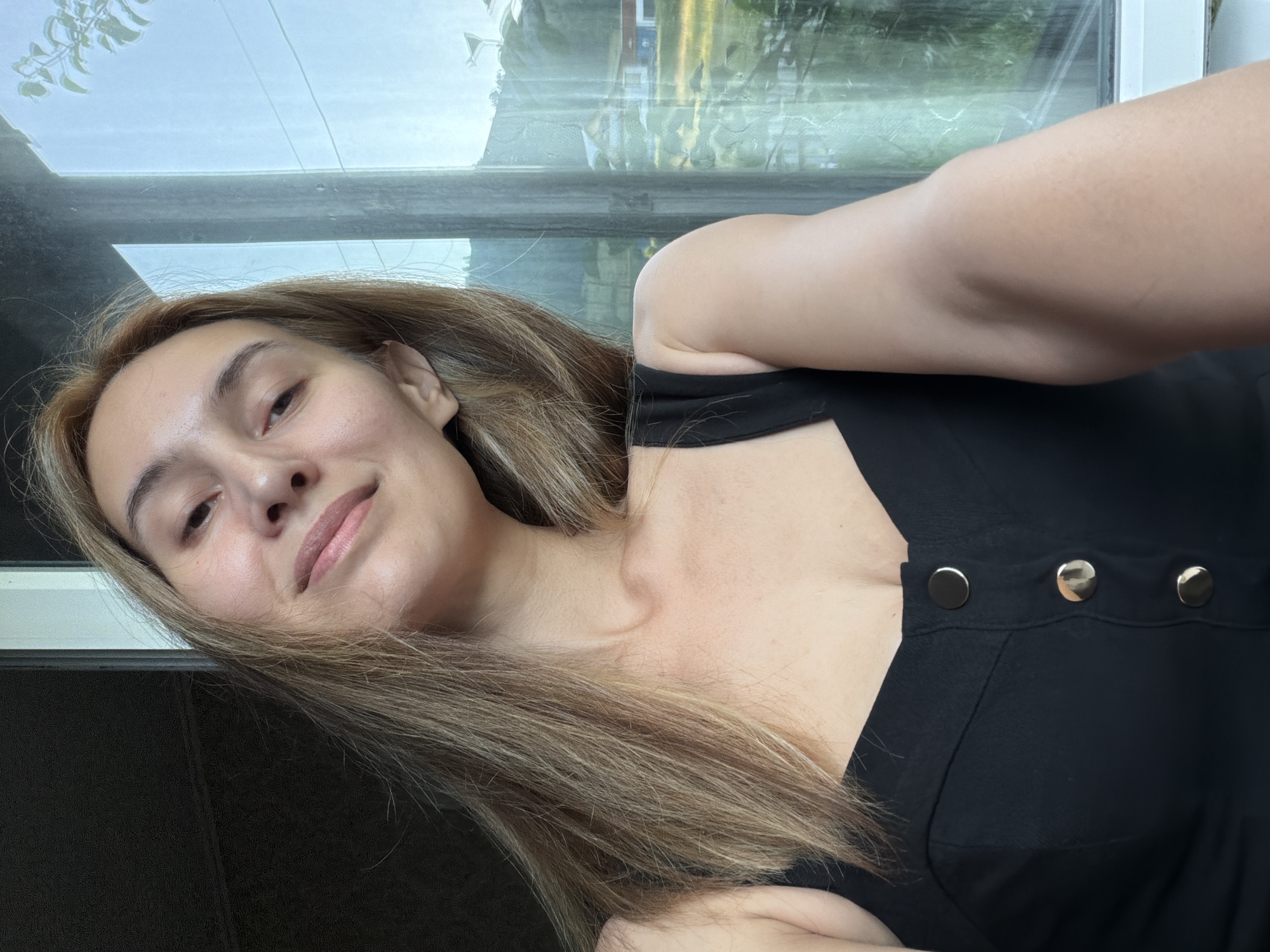 A young woman with long, light brown hair is shown in a selfie taken indoors, looking towards a window that overlooks a rural landscape. She is wearing a black sleeveless top with three decorative gold buttons. Her head is tilted slightly, and she has a subtle smile. The background outside the window shows trees, a house, and hills under a bright sky. The lighting suggests it is daytime, and the environment appears to be a residential area in the countryside.