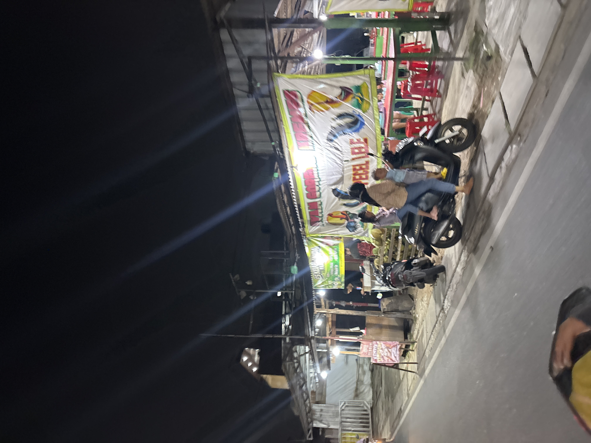 The image depicts a nighttime scene in South Tangerang, Indonesia, likely on a street or sidewalk. The primary subjects are people and motorcycles, engaged in various activities. In the foreground, a person is seated on a motorcycle. Behind them, a banner with colorful illustrations and text is prominently displayed, likely advertising a food stall or business. Several red chairs are visible, suggesting a seating area for customers.
The lighting indicates it's nighttime, with artificial lights illuminating the scene and casting shadows. The overall atmosphere appears to be that of a bustling street market or food vendor area, common in many parts of Indonesia. The presence of multiple motorcycles suggests transportation and perhaps a gathering of people.
One of the banners, though partially obscured and difficult to read completely due to the image's angle and resolution, appears to contain Indonesian text. Phrases like "AYAM GORENG" and "PECEL LELE" might be discernible, which are popular Indonesian dishes. This reinforces the idea of a food establishment.
Other details include what appear to be some stacked goods, possibly produce like coconuts, and the general structure of outdoor stalls or shops. The weather appears to be clear, as there are no visible signs of rain. The scene captures a slice of everyday life in an Indonesian urban or semi-urban environment at night.