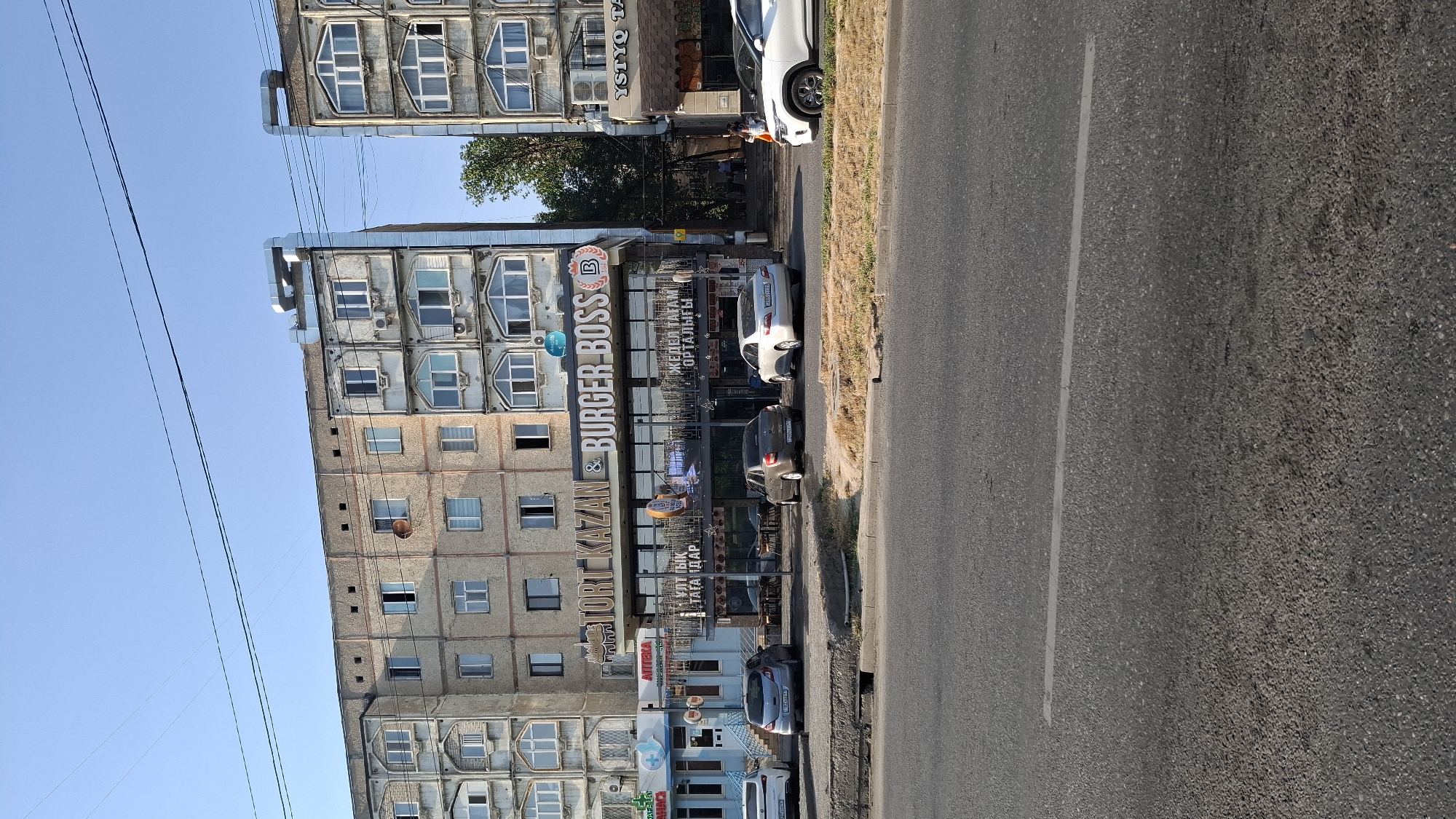 The image shows a street scene in the city of Shymkent, Kazakhstan, with a focus on a building housing a "TORT KAZAN & BURGER BOSS" establishment. The building is a multi-story apartment complex with a commercial ground floor. Several cars are parked on the street in front of the businesses. The weather appears to be clear and sunny. The architecture is typical of Soviet-era construction. The ground floor features glass storefronts and signage in what appears to be Cyrillic or a similar alphabet. One car is a white SUV parked on the right side of the street. Other cars visible are a silver sedan, a dark gray sedan, and a light gray hatchback. A person is seen walking on the sidewalk to the right of the Burger Boss establishment. A blue pharmacy with a red cross emblem is visible to the left of Burger Boss. The sky is clear blue with visible power lines overhead. The street in the foreground is asphalt with a painted white line.