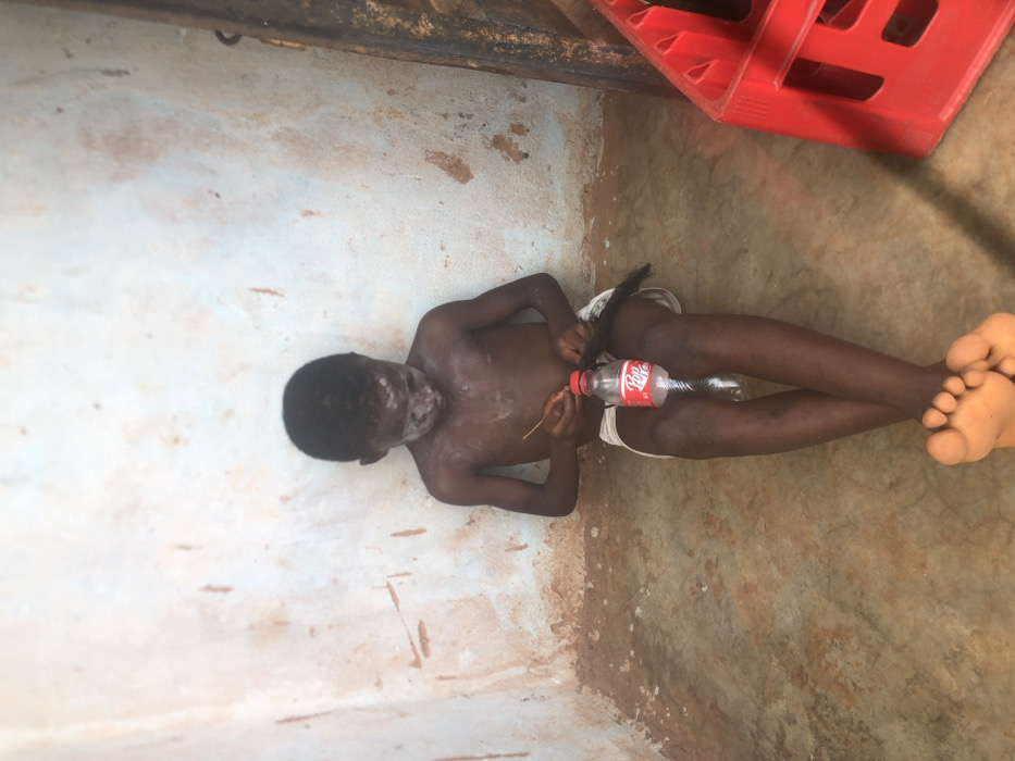 Inside a dilapidated structure in Ukwu-Abwa, Nigeria, a young, dark-skinned male child is seated on a mottled brown floor, facing forward with his gaze directed downwards. He is shirtless, wearing light-colored shorts, and has a white powdery substance applied to his face and upper chest. His bare feet are visible, crossed at the ankles. Between his legs rests a clear plastic Coca-Cola "Pop" bottle with a red cap, appearing to be cut or modified at the base. The child uses a thin, dark stick-like object in his right hand to manipulate a dark, fibrous material held in his left hand, positioned near the modified bottle. The background consists of a worn wall, painted with light blue and white patches over a reddish-brown base, showing significant discoloration and peeling, especially near the floor line. To the right of the child, a portion of a red plastic crate, containing dark bottles, is visible.