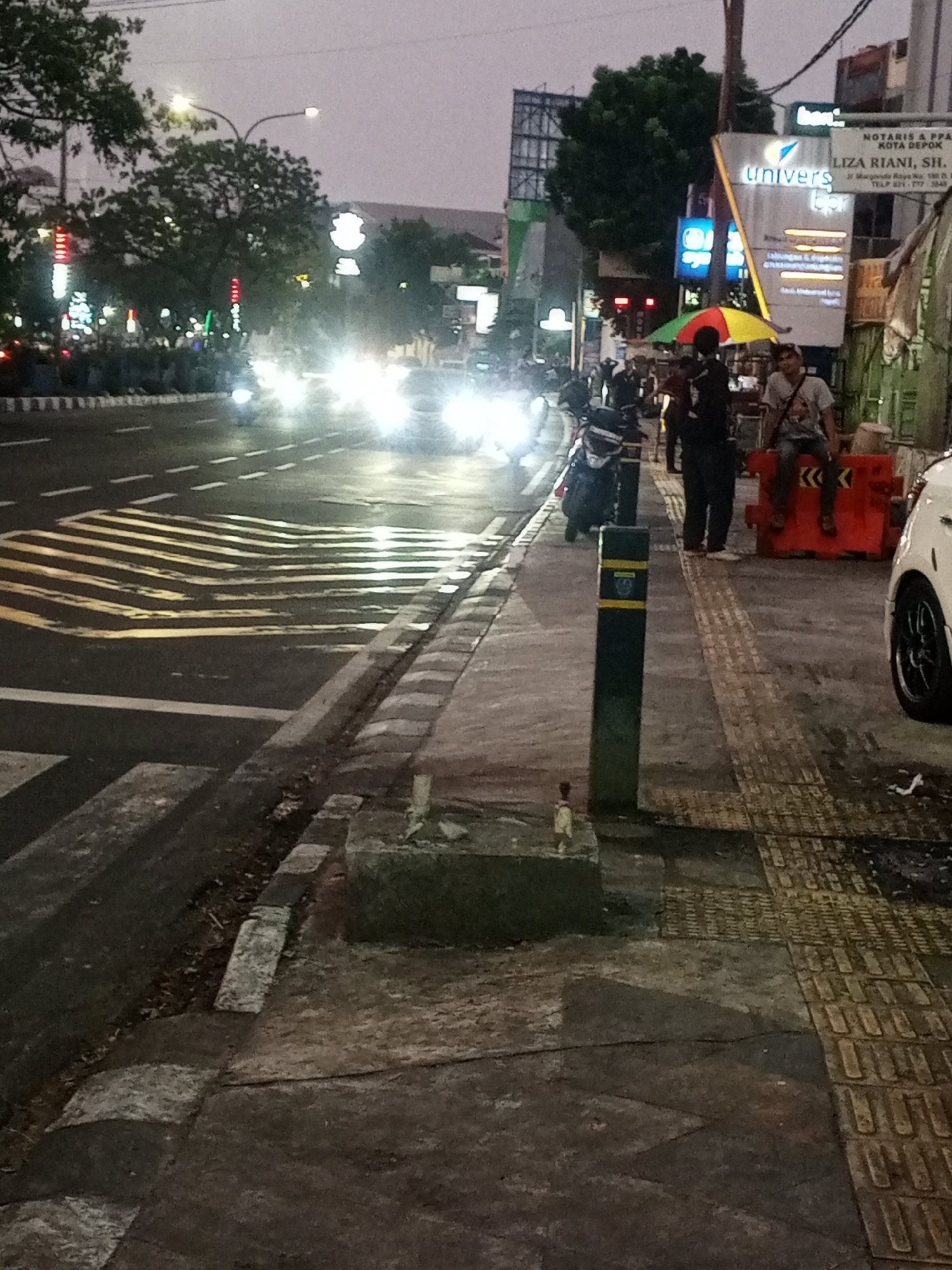 The image depicts a street scene at dusk in Depok, Indonesia, likely near a commercial area. The foreground shows a textured sidewalk with raised tactile paving, a dark bollard with a yellow band, and a low concrete barrier. A white car is partially visible on the right. The street in the midground features yellow and white road markings, including a speed bump, and is illuminated by the headlights of oncoming traffic, suggesting it is a busy road. Several motorcycles are parked or moving on the street and sidewalk. In the background, buildings with illuminated signage are visible, including "Univers," and advertisements for legal services. There are also trees and streetlights lining the road. A few individuals are present on the sidewalk; two men are seated on an orange barrier, and others are standing near a colorful umbrella. The overall atmosphere is that of a bustling urban environment at the end of the day.