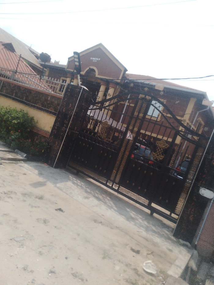The image depicts the exterior of a property in Iranla, Nigeria, captured from an unpaved dirt road surface under daylight conditions. The scene is dominated by a large, ornate, double-leaf metal gate, primarily dark in color with gold-colored decorative elements. The gate features solid panels at its lower section and intricate scrollwork and decorative motifs in its upper half. It is flanked by dark, patterned gate posts. A small, triangular red and white sign is affixed to the right leaf of the gate.
Behind the gate, a multi-story building is visible, characterized by a prominent gabled section at the front, painted dark reddish-brown with lighter trim. The words "BY HIS GRACE HOTEL" are legible in light lettering on this gable. The building's facade is dark reddish-brown with lighter, possibly cream-colored, architectural details such as columns or pilasters. Windows with dark frames are visible. A dark-colored vehicle is partially discernible parked inside the property, beyond the gate. The roof appears to be tiled in a reddish-brown color.
To the left of the gate, a perimeter wall constructed of dark, possibly stone or concrete blocks, extends, topped with a dark barrier. Green shrubbery with scattered red flowers grows along the base of this wall. A black solar street light is mounted on a pole, positioned to the left of the gate structure and extending upwards. Overhead, utility wires traverse the upper portion of the frame against a bright, hazy sky. The foreground road surface is light-colored dirt with minor debris. No individuals are visible in the scene.