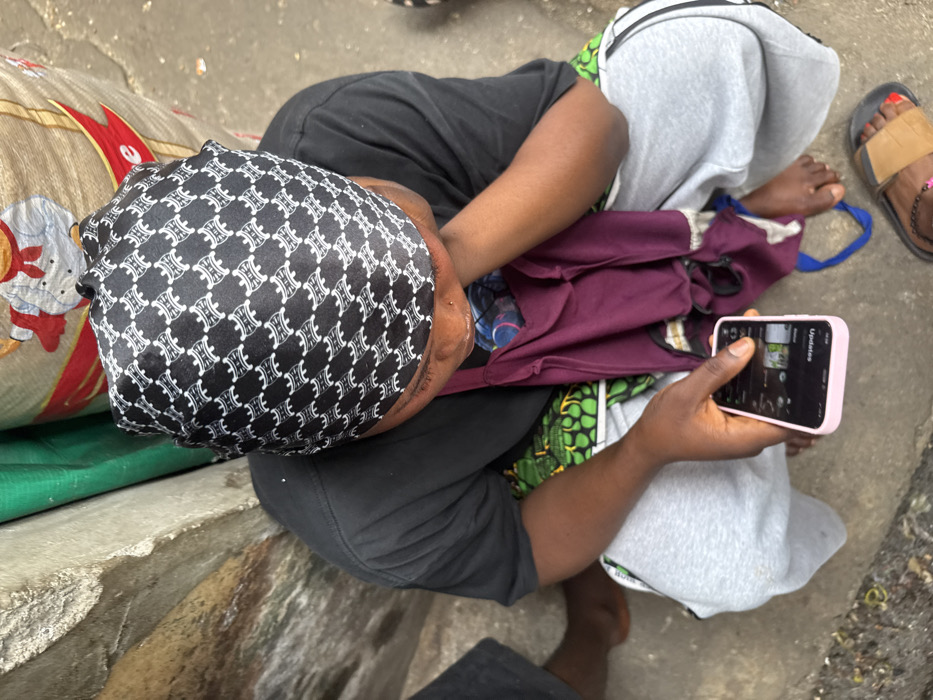 A person sits on the ground, wearing a patterned head covering, a black t-shirt, and gray sweatpants. They are holding a smartphone with a pink case. A maroon shirt is partially visible on their lap. A sandal with a red-painted toe is seen near the right edge. There is a patterned bag to the left. The ground is concrete and somewhat dirty.