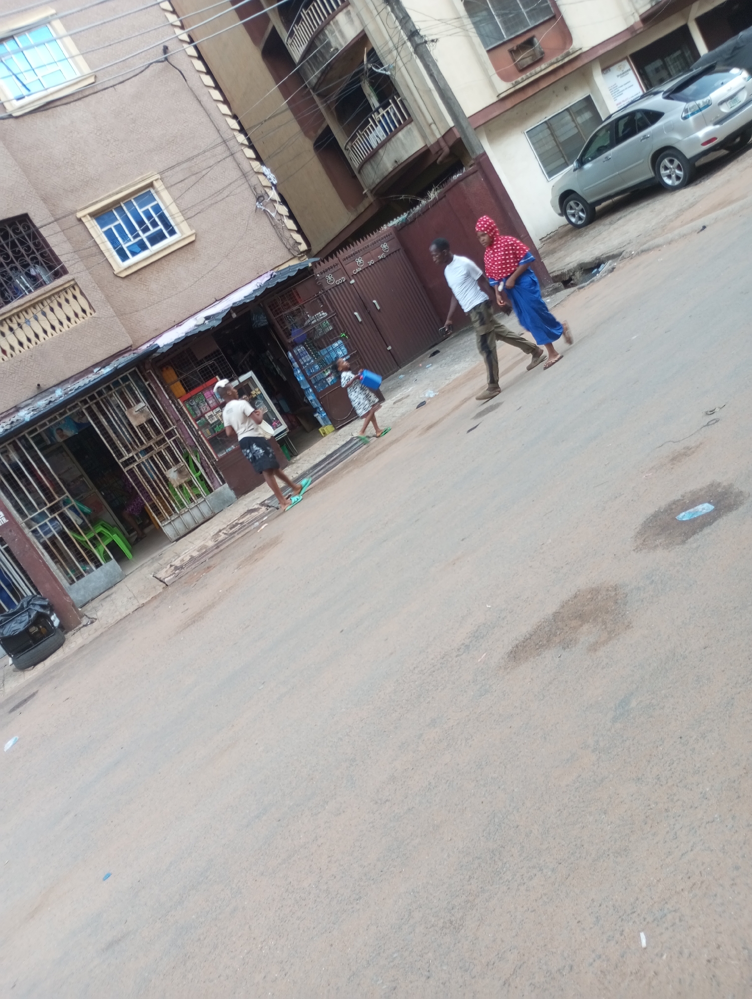 This image captures a street scene in Onitsha, Nigeria, likely during the daytime. The perspective is slightly angled, looking down the street.
The scene depicts a busy urban or semi-urban environment lined with multi-story buildings. On the left, a brown building with patterned walls features multiple windows, some with blue-tinted glass, and balconies with decorative railings. Its ground floor is occupied by several shops, enclosed with metal gates and grilles, displaying various goods. Green plastic chairs are visible inside one shop entrance. A black tire with a bag on it rests on the pavement in the bottom-left corner.
On the right side of the street, another building, possibly lighter in color, has a silver Lexus RX series SUV parked in front of it. A sign with the word "DIAMOND" is visible on this building. Overhead, numerous electrical wires crisscross the sky.
Several people are engaged in daily activities on the street. A man in a white t-shirt and camouflage pants walks away from the camera, accompanied by a woman in a red and white patterned top, a blue skirt/dress, and a red head covering. Both are wearing flip-flops. Further left and closer to the foreground, another man in a white t-shirt and dark shorts, also in green flip-flops, walks past the shops. A child in a light-colored dress, carrying a blue bucket, is partially visible behind him. People appear to be going about their errands or daily commutes.
The asphalt road shows signs of wear, with some dirt patches and small debris. The light suggests it is daytime, possibly late morning or early afternoon, with diffuse light rather than direct sunlight. Visible text includes "DIAMOND" on a sign and partially obscured text on a metal gate reading "CO-OP" and "NO 88".
