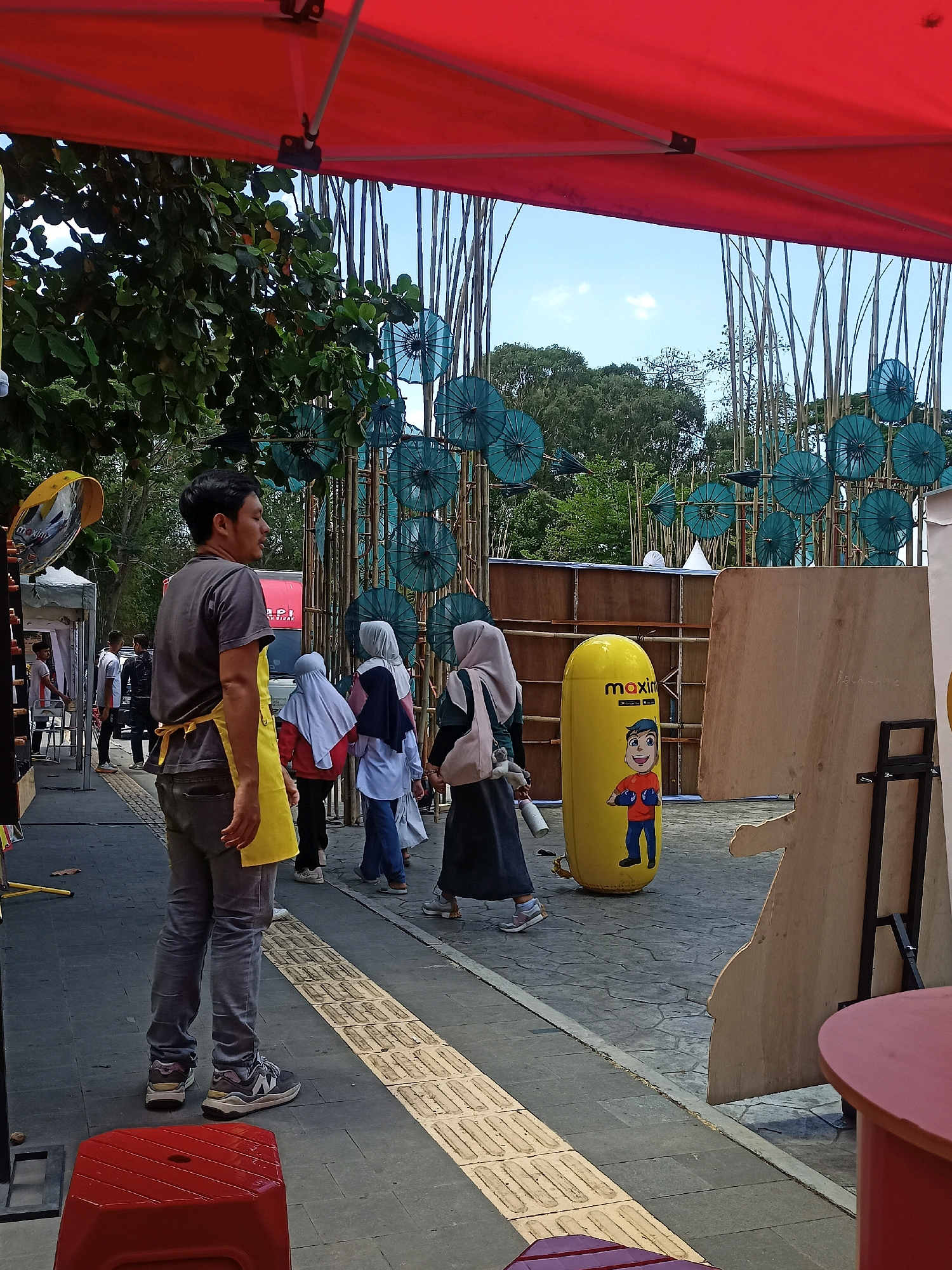 The image captures a scene outdoors under a large red tent, possibly at an event in Surakarta, Indonesia. In the foreground, a man wearing a grey t-shirt and grey jeans stands facing away from the camera, his head turned to the side. He has a yellow apron tied around his waist. A red plastic stool is visible in the lower left corner.
Behind the man, several people are walking. One person, seen from the back, wears a light blue hijab and a dark blue top with a white shawl. To their left, another person wears a light blue hijab and a red top. Further back, more individuals are visible, some wearing hijabs.
The environment features a paved walkway with a tactile paving strip. In the background, there is a decorative structure made of bamboo poles adorned with numerous teal-colored, umbrella-like objects. These are likely traditional paper umbrellas, a common craft in Indonesia. There is also a tall, yellow inflatable punching bag with a cartoon character printed on it, featuring the text "maxim." A large, unfinished wooden cutout in the shape of a person stands to the right. Trees and a clear blue sky are visible beyond the bamboo structure. A red tent canopy is stretched across the top of the image, providing shade. The scene appears to be a public space, possibly a street or a market area during an event.