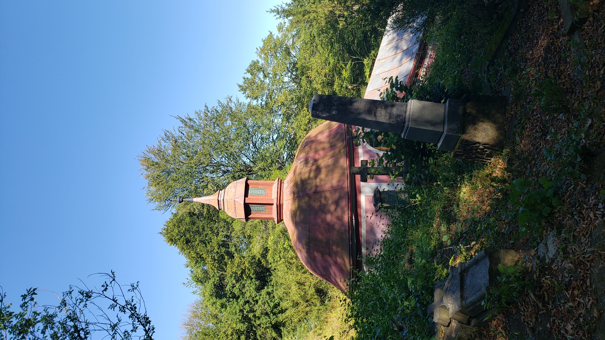 The image depicts an outdoor scene featuring a domed building with a spire, surrounded by lush green trees and vegetation. In the foreground, there are several tombstones and what appear to be graves, suggesting a cemetery. The building's dome is a rusty red color, and the spire has a smaller, more ornate structure at its peak with windows. The sky is a clear, bright blue. The lighting suggests it is daytime, with sunlight casting shadows on the building and tombstones. This scene is located in the Old Cemetery (Starý cintorín) in Prešov, Slovakia.