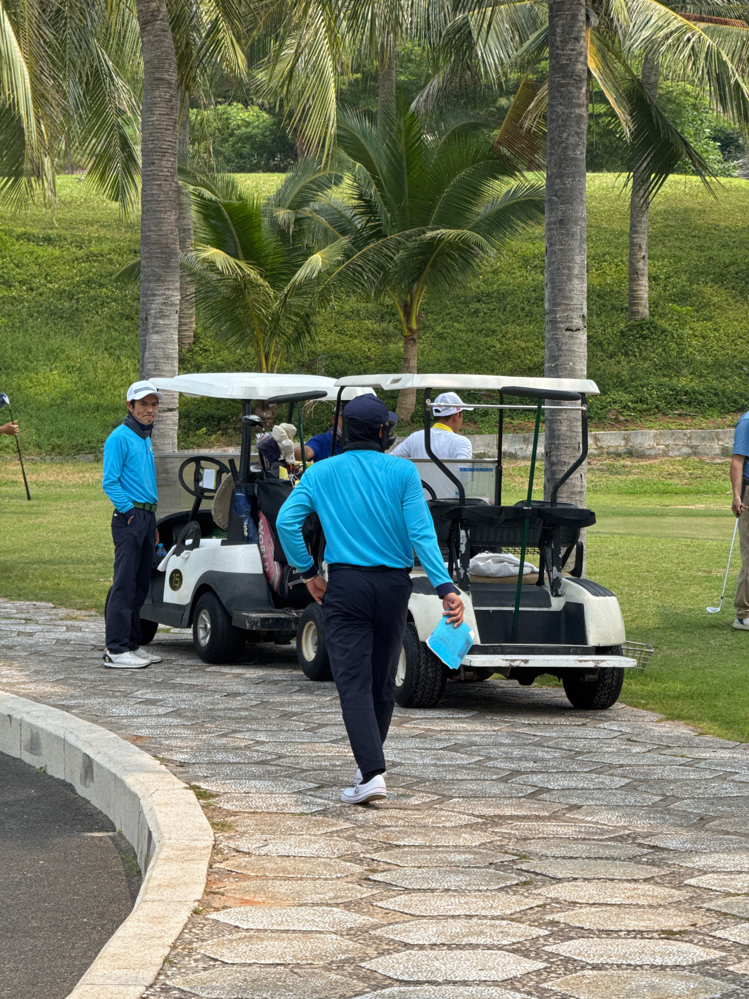 The image depicts a scene at a golf course in Phu Thuy, Vietnam, on a cloudy day. In the foreground, a person wearing a blue long-sleeved shirt and dark pants walks away from the viewer, holding a blue sheet of paper. To the left of this person is a white golf cart with a canopy, parked on a paved pathway. Two individuals are standing near the golf cart; one in a blue long-sleeved shirt and dark pants, and another in a white cap and a yellow shirt is seated in the golf cart. Behind the golf cart, palm trees and lush green grass are visible, characteristic of the tropical environment of Vietnam. The pathway is made of interlocking stone slabs, with a curb separating it from the grassy area. In the background, more palm trees and verdant landscape suggest the location is a well-maintained golf resort.