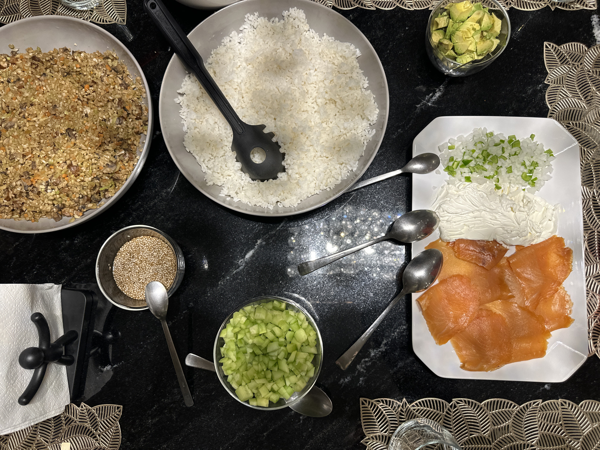 The image shows a tabletop arrangement of various ingredients, potentially for a build-your-own sushi or similar dish.
From left to right, there is: a gray bowl with what appears to be cooked and chopped mushrooms and other vegetables, a bowl of cooked white rice with a black serving spoon, a glass bowl of diced avocado, a white plate with smoked salmon, a dollop of cream cheese, and diced onions and green peppers. Additional elements include a bowl of sesame seeds, a glass bowl with diced cucumber, a black napkin holder, and several small spoons. The items are positioned on a dark surface, possibly a marble countertop.
There is nothing to indicate that the image was taken in Mexico City.