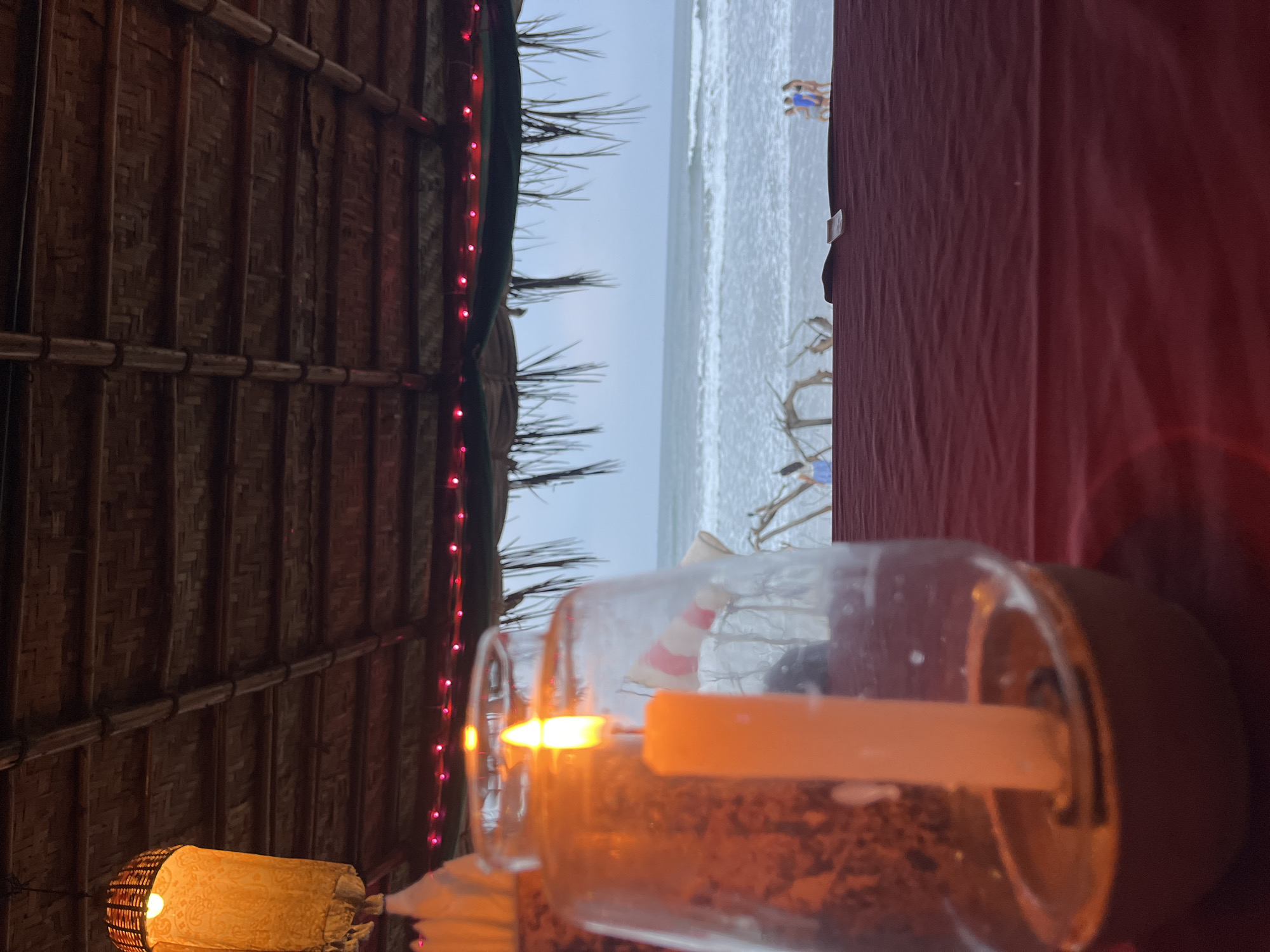 The image shows a beach scene viewed from an indoor or sheltered location, likely a restaurant or hut. The beach is in the background with visible people near the water. In the foreground, there is a lit candle inside a glass container, providing ambient light. The structure overhead appears to be thatched or made of woven material. The side of the scene is partially obscured by a red cloth. The location is Ashvem, India.