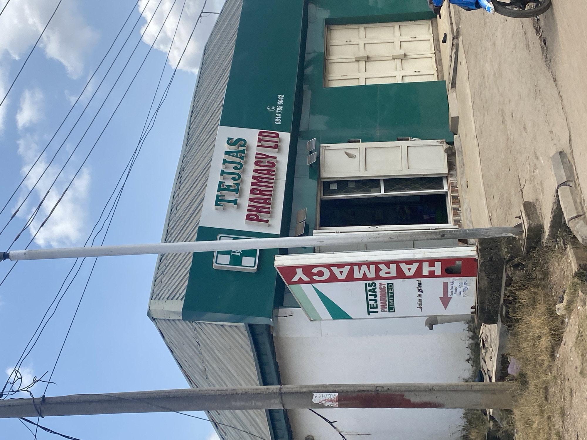 The image shows the exterior of a pharmacy in Ado, Nigeria. The building is painted green and white, with a prominent sign that reads "TEJJAS PHARMACY LTD". A smaller sign on the side of the building also says "TEJJAS PHARMACY LIMITED" with the text "...balm in gilead" underneath. There is a phone number visible on the main sign, "0814 788 6042". The pharmacy has a glass door and two large, beige-colored doors that appear to be folded open. In front of the pharmacy, there are two utility poles, one made of concrete and the other a thinner white pole. Overhead, electrical wires are visible against a blue sky with scattered clouds. A blue motorcycle is parked to the right of the building, and the ground in front of the pharmacy is a paved surface with some patches of dry grass and debris.
