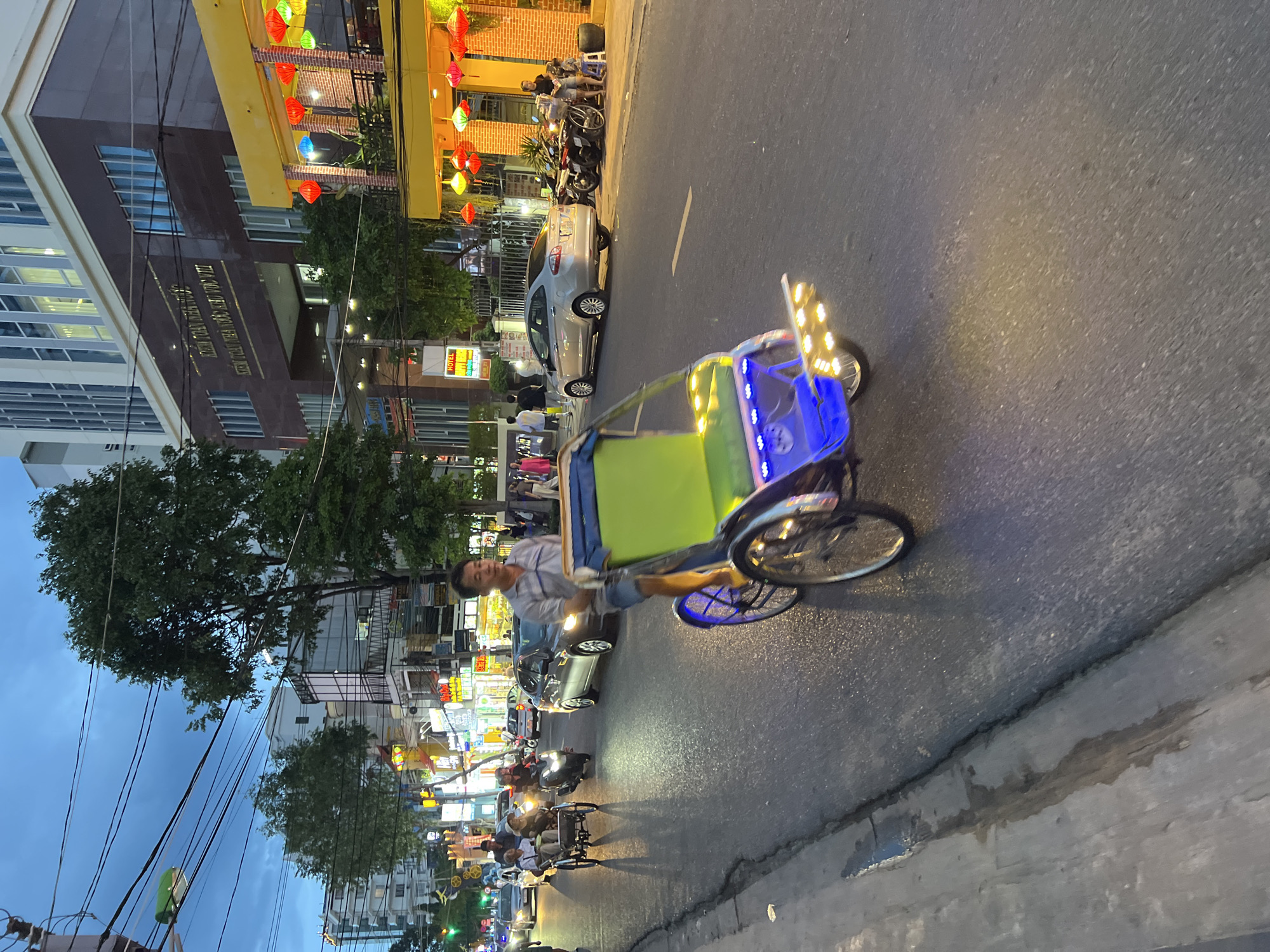 The image depicts a street scene in Nha Trang, Vietnam, at dusk. A man is riding a cyclopousse, a three-wheeled bicycle taxi, with a green seat and blue lights on the back. The cyclopousse is in the foreground and is moving along the right side of a paved road. In the background, there are several cars, including a silver sedan, and a motorbike. Buildings line the street, some with bright signage and illuminated windows. One building, in the mid-ground, has the words "KIEM TOAN NHA NUOC" visible on its facade. Red and yellow lanterns hang from the awning of a building on the right side of the street, suggesting a commercial area, possibly a restaurant or shop. The sky is a deep blue, indicating the transition from day to night, and power lines are visible crisscrossing overhead. The overall impression is of a busy urban environment during the evening hours.