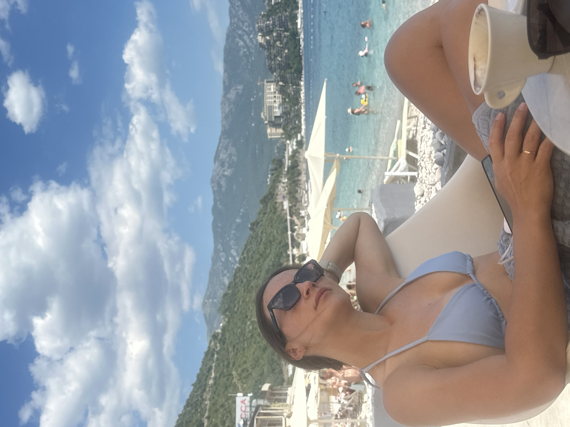 A woman with dark hair and sunglasses is lying on a lounge chair at a beach, facing a scenic view of the sea and mountains. She is wearing a light blue bikini. Her left arm is resting behind her head, and her right hand is holding a grey towel with a ring on her finger. A white cup is visible next to her.
The background shows a clear blue sky with fluffy white clouds. The sea is a vibrant turquoise color with several people swimming and wading in it. In the distance, a mountainous landscape is covered in lush green vegetation, and buildings are nestled along the coastline. A white umbrella is set up on the beach.
The scene suggests a sunny day, likely in the late morning or early afternoon given the bright sunlight. The overall mood appears relaxed and vacation-like, with the woman enjoying the beach setting. The location is contextually identified as Budva, Montenegro. No discernible text is visible in the image.