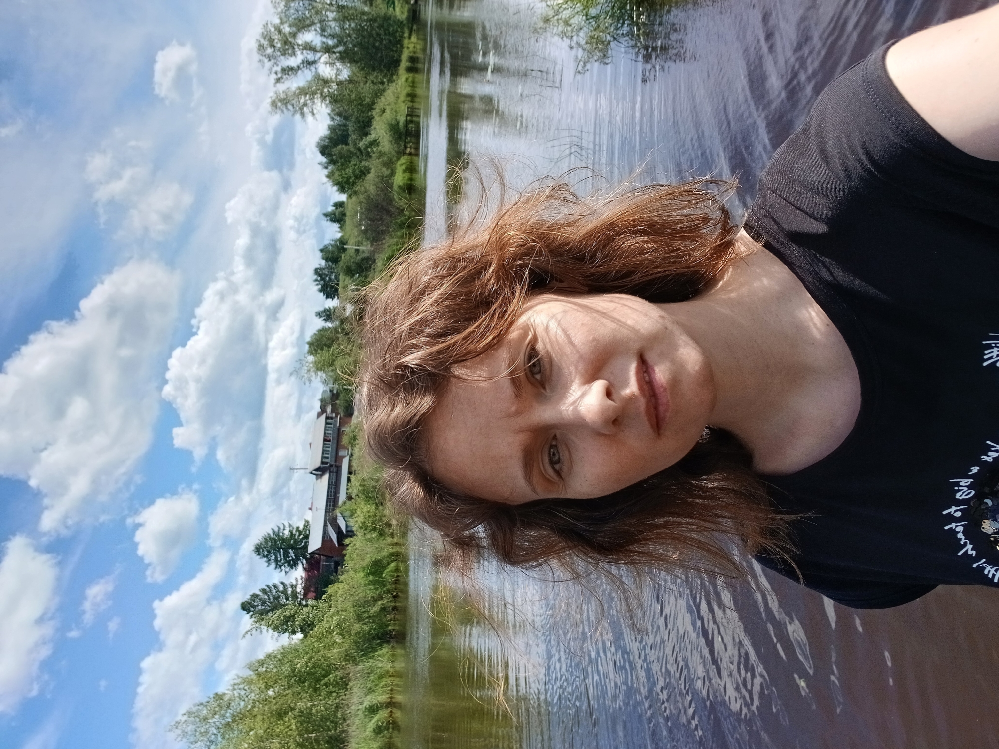 A selfie is taken outdoors on a bright, sunny day with fluffy white clouds scattered across a blue sky. The scene is dominated by a calm body of water that reflects the sky and surrounding greenery. On the far bank of the water, there is a line of lush green trees and bushes, with a few buildings visible amidst the foliage. The buildings appear to be red-roofed and have a rustic, possibly residential, feel.
In the foreground, a person with light brown, wavy hair is looking towards the camera. Their face is turned slightly, and they have freckles across their nose and cheeks. The person is wearing a black t-shirt. The image is oriented with the horizon line running vertically through the frame, suggesting the photo was taken at an angle. The overall mood appears relaxed and natural, possibly indicating a leisurely moment by the water. The presence of trees, water, and open sky suggests a natural, perhaps rural or semi-rural, setting. The text on the person's t-shirt is partially visible and appears to be in a white, handwritten font.