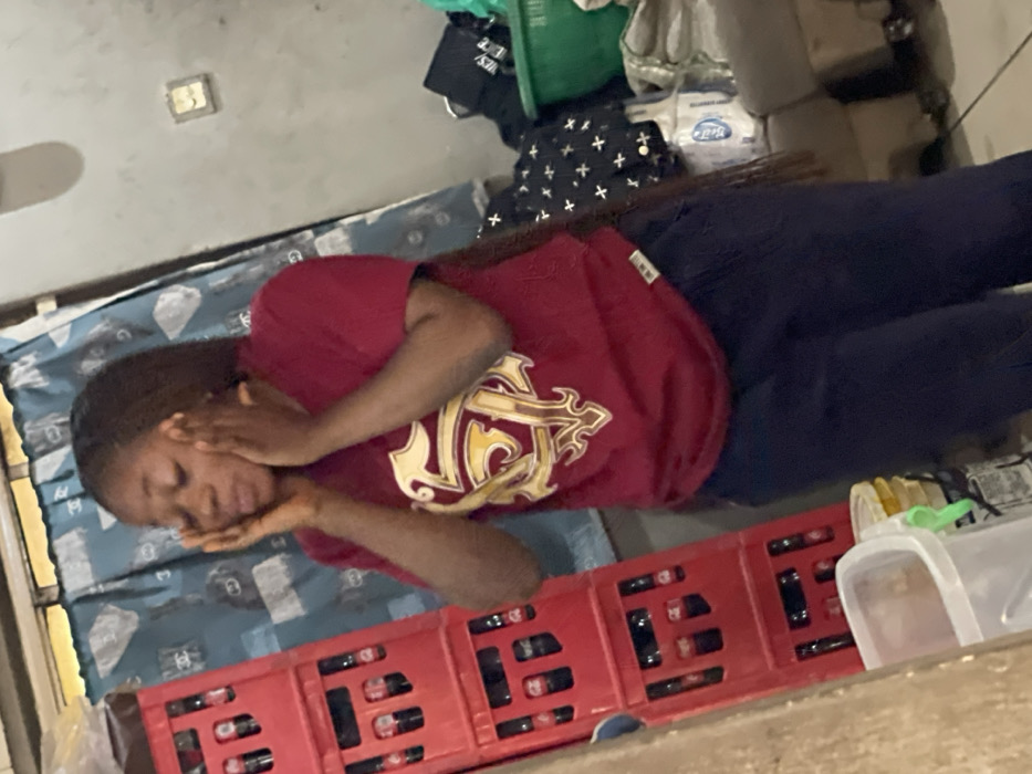 A young woman lies on a bed, her head propped on her hands. She's wearing a maroon t-shirt with a large gold crest. The bed has a blue and white patterned cover. Next to her is a red plastic crate filled with dark bottles. In the background are assorted items including a green basket, bags with white crosses, and white boxes. A light switch plate is on the wall behind the bed. She looks distressed.