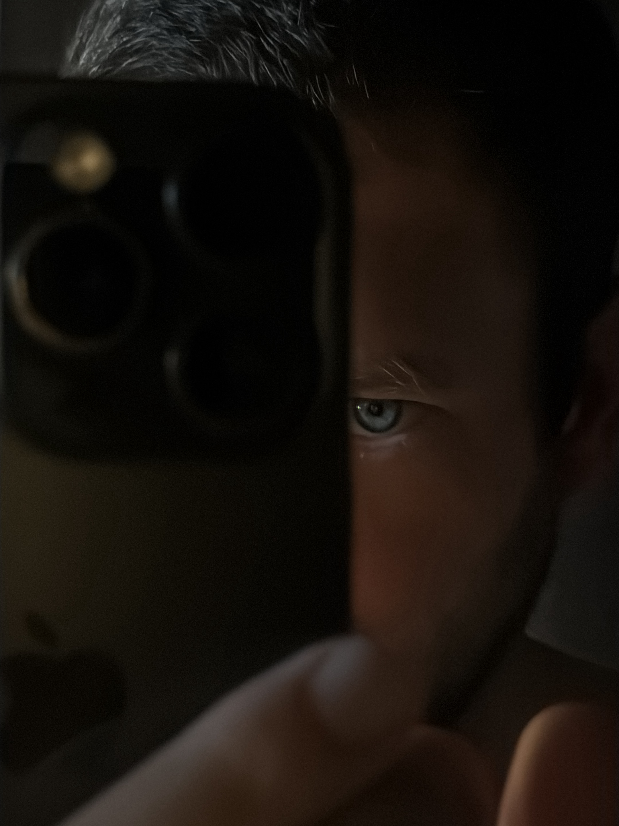 An extreme close-up shot captures a man's eye as he takes a selfie with a smartphone. The smartphone is held in the foreground, partially obscuring the man's face, with its camera lenses visible. The man's eye is the primary focal point, showcasing a bright blue iris with a subtle reflection. The background is dark and indistinct, suggesting an indoor setting. The lighting is dramatic, casting shadows and highlighting the textures of the man's skin and hair. The image has a gritty, candid feel. There is no indication of the image being taken in Milan, Italy.