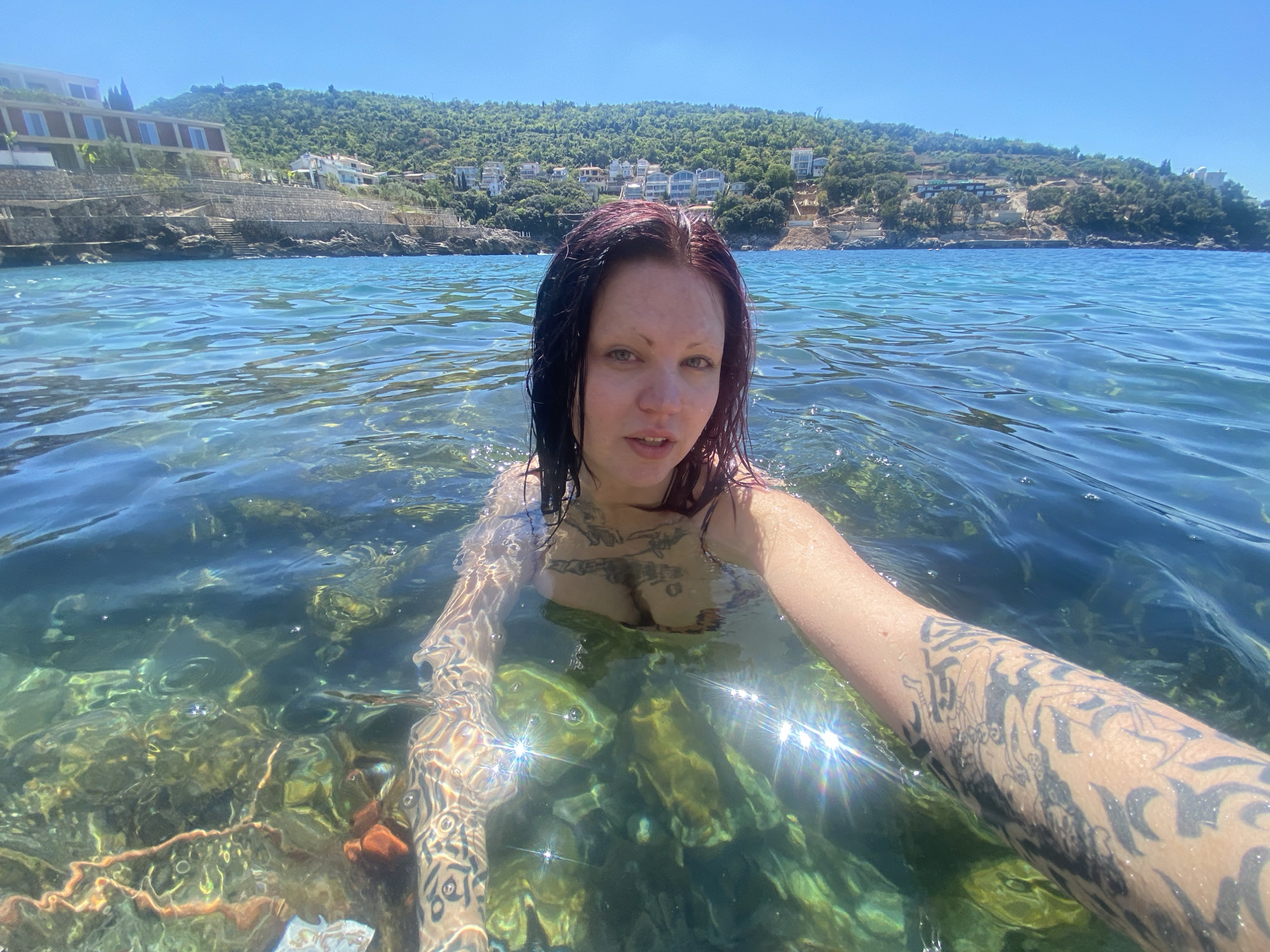 A woman with dark hair and tattoos swims in clear, turquoise water. She is holding the camera for a selfie. Her tattoos are visible on her arm and chest. The seabed is visible through the clear water, with rocks and seaweed. In the background is a hill covered in green vegetation and buildings, a seaside resort area. The location is the Adriatic Sea. The area in the city of Kunje, Montenegro. It is daytime, and the sky is clear. The weather is sunny.