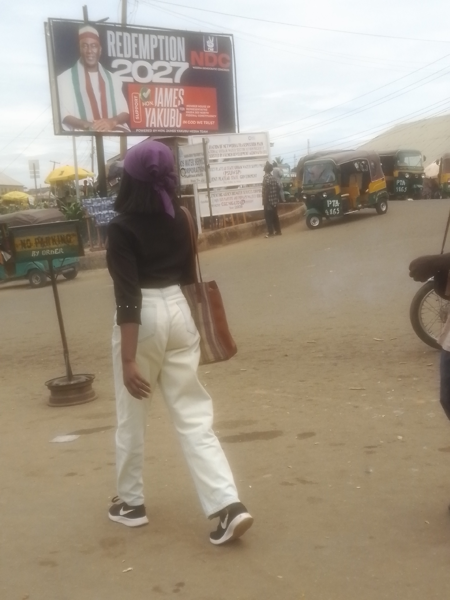 In Gwong, Nigeria, a young woman walks away from the camera, her back to the viewer. She wears a black top with pearl-like embellishments on the sleeves and loose-fitting, light-colored pants. A purple headscarf covers her hair, and she carries a striped tote bag over her shoulder. Her black Nike sneakers are visible as she walks on a dusty, paved road.
In the background, a large billboard displays "REDEMPTION 2027" and the logo of the Nigeria Democratic Congress. The billboard features a portrait of a man in traditional attire and text identifying him as "HON. JAMES YAKUBU," running for House of Representatives for the Bassa 300 North Federal Constituency. Another sign reads "NO PARKING BY ORDER."
Several auto-rickshaws, known as "keke napeps," are present on the road, one with the license plate "PTA 4165" and another with "PTA 9165." Other signs in the background are partially obscured but appear to be official notices related to water services and government agencies. The scene suggests a public area, likely a market or street intersection, possibly in the morning or afternoon given the lighting. The overall atmosphere seems ordinary and transactional, typical of a busy urban or semi-urban environment in Nigeria.