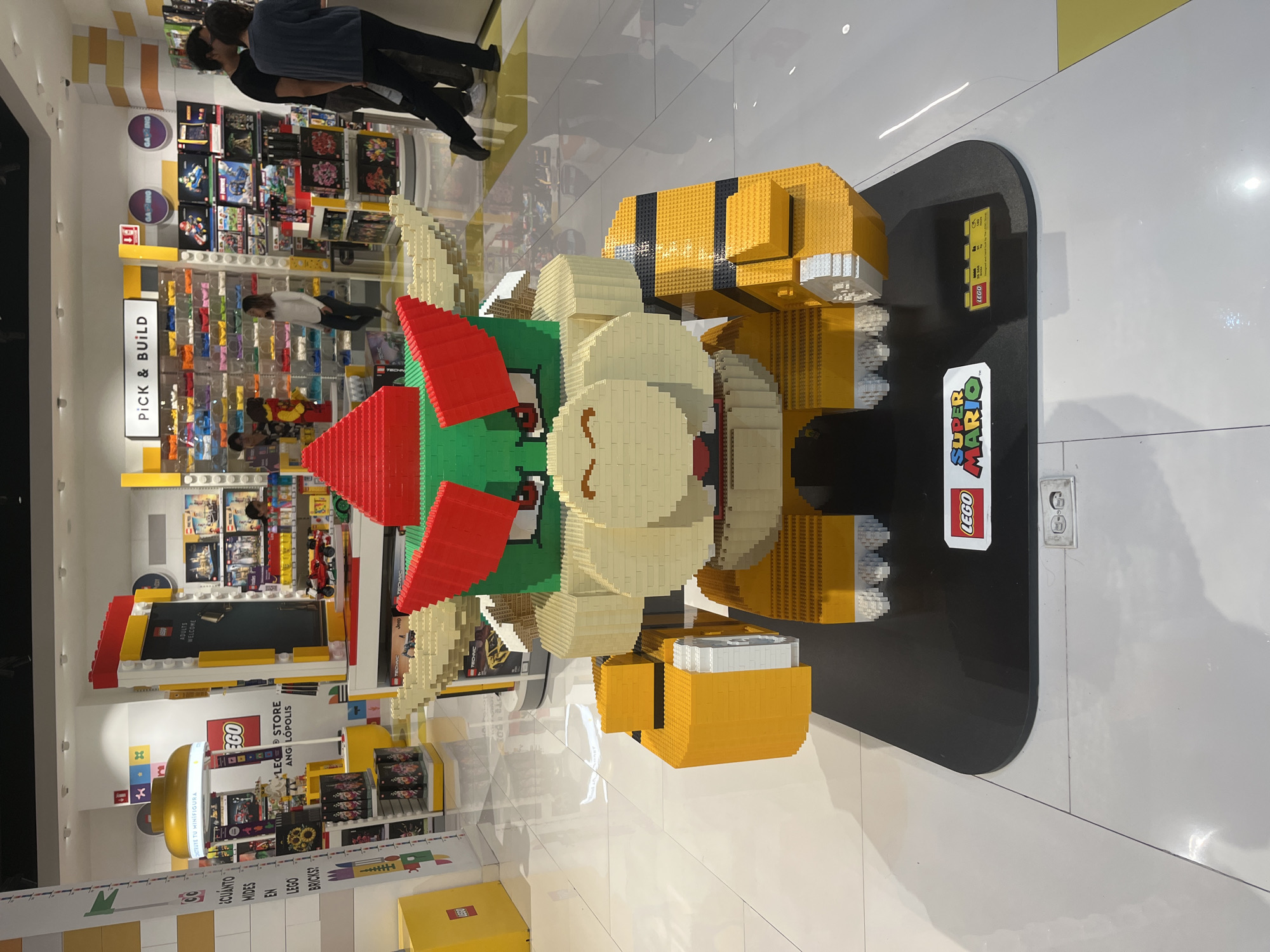 Here is a description of the image:
The image shows a life-size Lego sculpture of Bowser, the main antagonist from the Super Mario video game franchise, in a brightly lit Lego store. The sculpture is made entirely of Lego bricks and stands on a black platform with the "LEGO Super Mario" logo prominently displayed. An information card is next to the logo on the platform. In the background, the Lego store has shelves stocked with Lego sets, a "Pick & Build" section with bins of loose Lego bricks, and other displays. People are browsing the Lego products. The store appears to be located in Angelópolis, a district of Puebla, Mexico, judging by the signage on the store's wall.