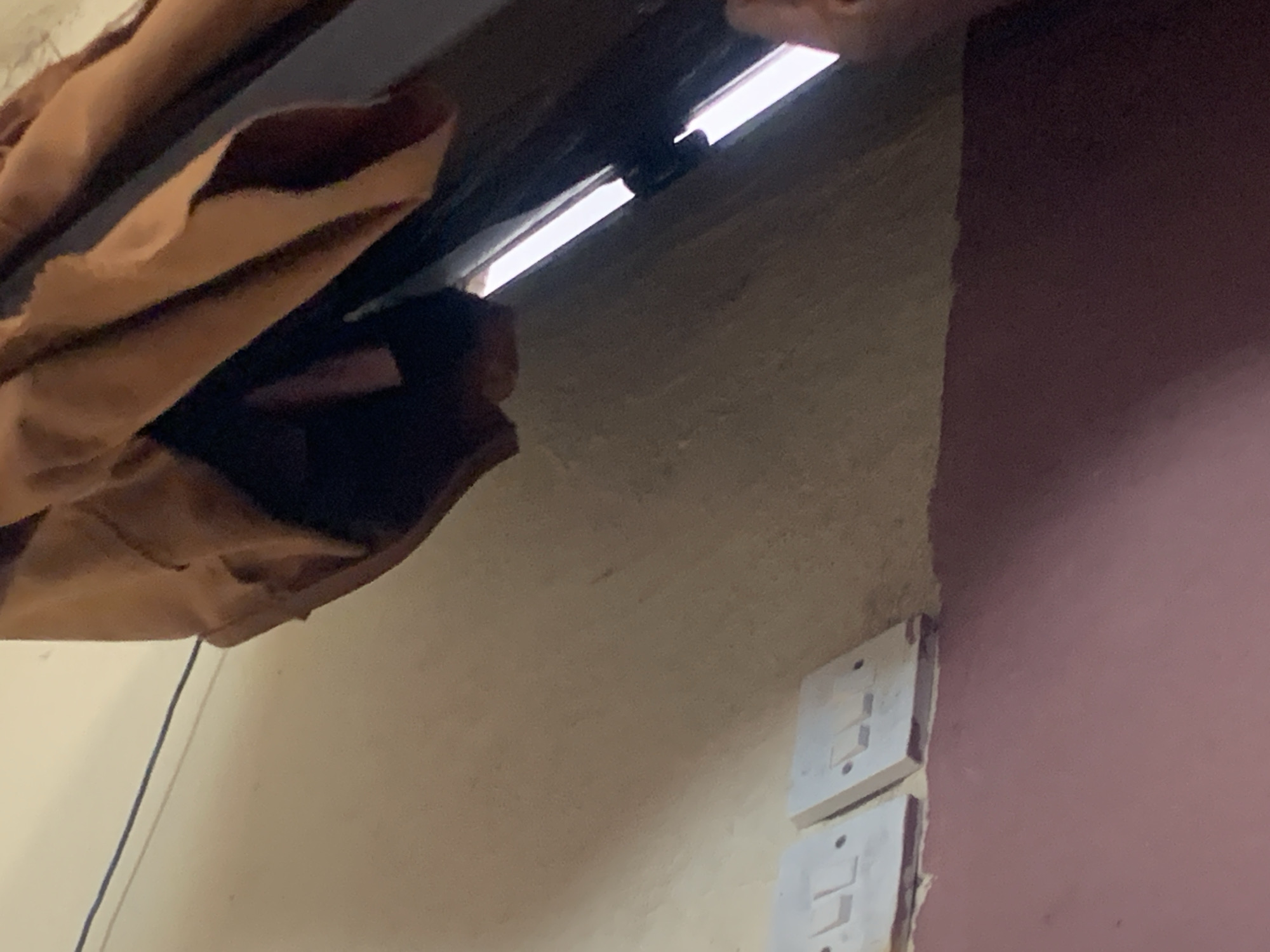 The image depicts an interior scene, likely a wall with electrical outlets. Two white electrical outlets are visible on the left side of the frame, mounted on a beige-colored wall. The outlets appear to be standard two-gang switch plates. Above the outlets, a thin dark wire runs horizontally across the wall. To the right of the outlets, a large piece of fabric, appearing to be brown or rust-colored, is draped downwards. Part of a dark, reflective surface is visible behind the fabric, possibly a window or a dark wooden surface, with bright light emanating from behind it, suggesting a light source or opening. The lower portion of the image shows a transition from the beige wall to a dark pink or reddish-brown painted surface. The overall impression is of a functional, yet somewhat unfinished or cluttered, domestic space. Without additional context, it is impossible to pinpoint the exact location within Jauro Yinu, Nigeria. However, the presence of electrical outlets suggests an inhabited structure.