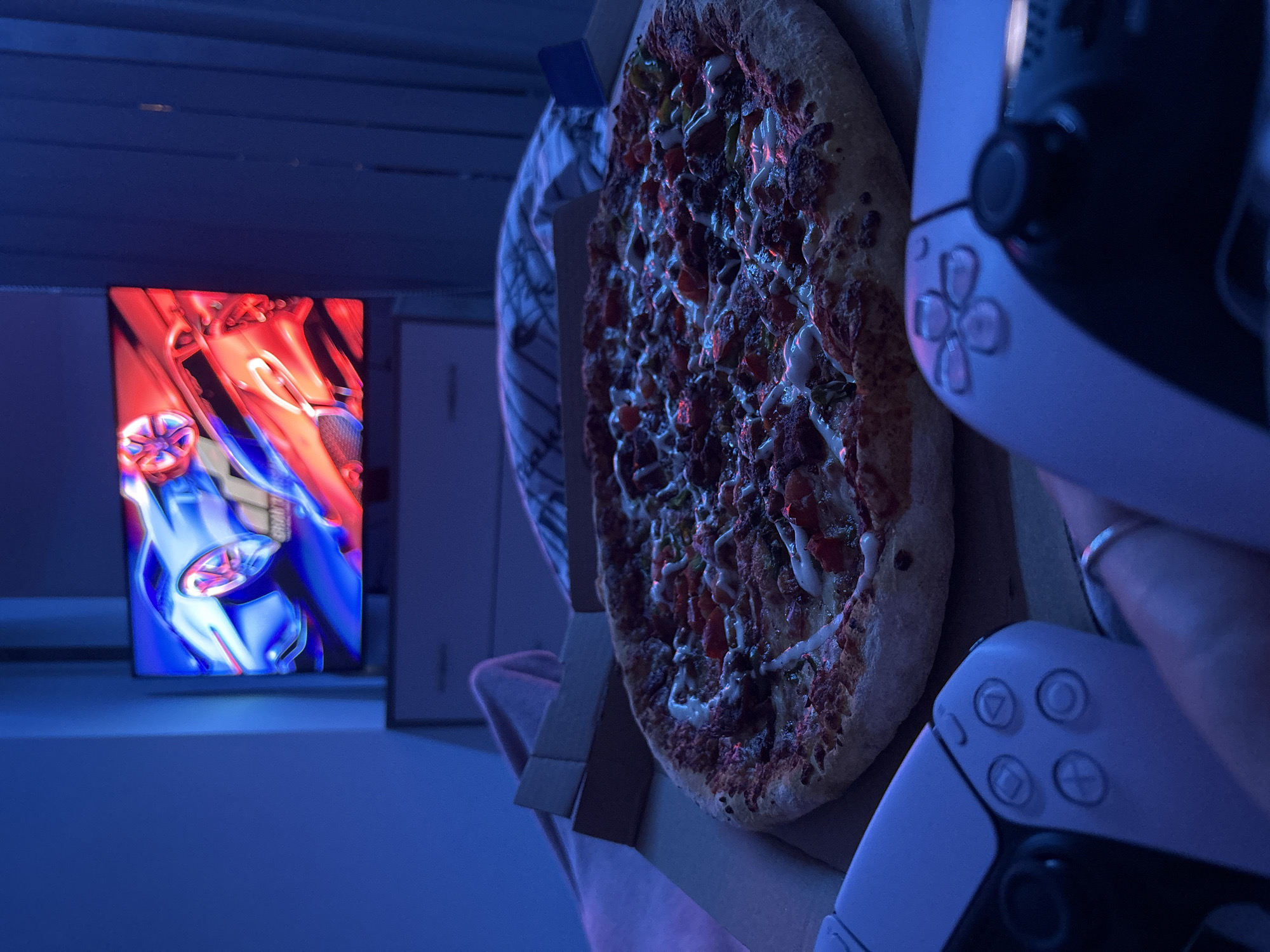 The image shows a lit display screen displaying an image of stylized robot or character. Adjacent to the screen is a pizza in a cardboard box. Next to the pizza box are two PlayStation 5 DualSense controllers. The scene is in a room with blue-toned lighting. The location is not discernible from the image, so it is not possible to identify if the location is in Sofia, Bulgaria.