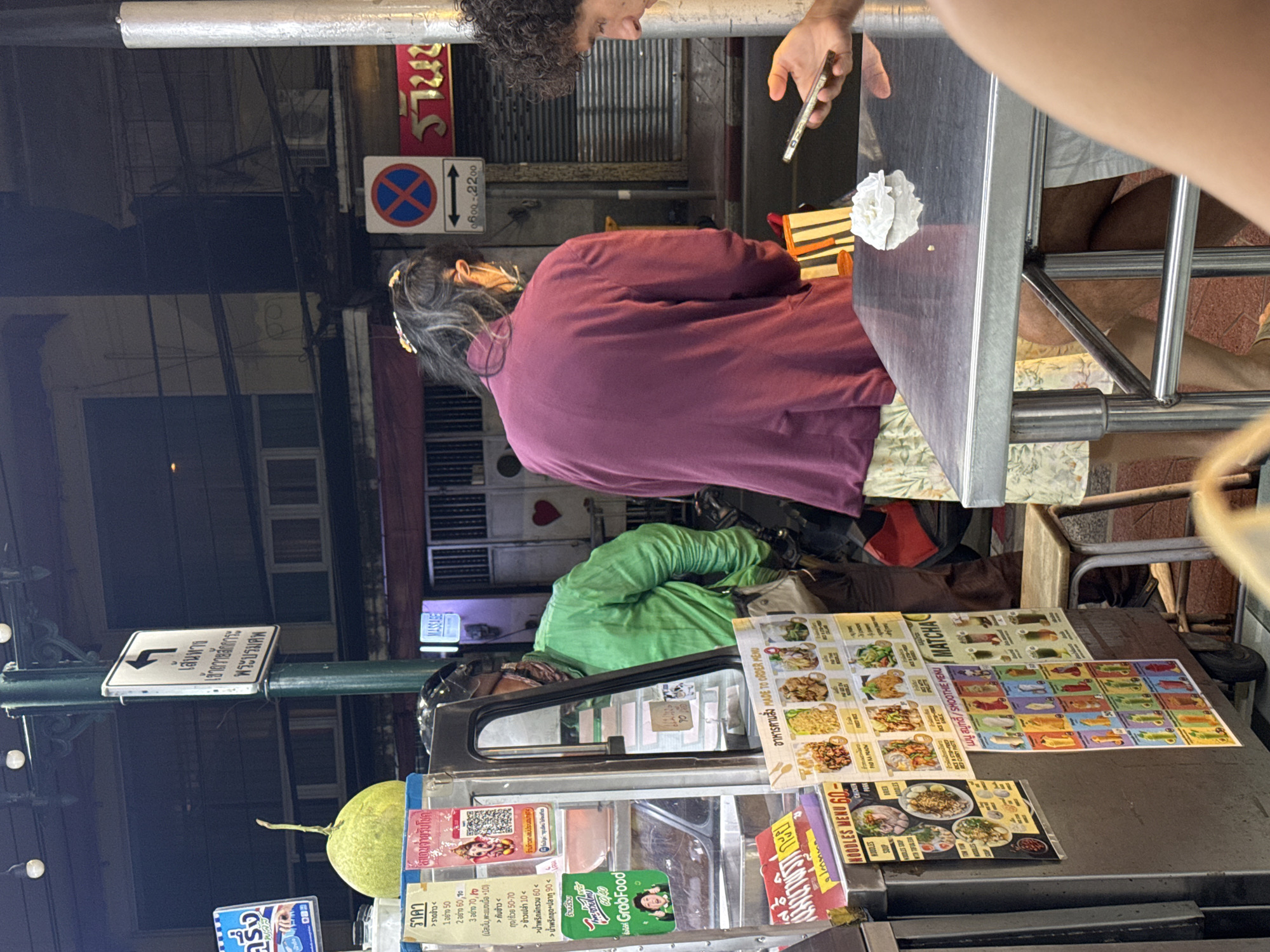 In Bangkok, Thailand, at night, a food stall is set up on a street. A person wearing a green shirt and a helmet stands behind the stall. A person wearing a dark purple jacket faces away from the camera. A third person with curly hair looks at their phone. The food stall has several signs displaying menus in Thai and English. The street appears to be lined with buildings and streetlights. The overall impression is a bustling street food scene.
