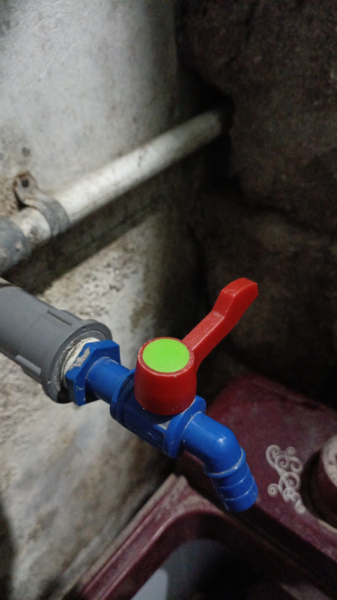 A close-up shot focuses on a blue PVC pipe connected to a red and green ball valve. The valve has a red handle with a green circular insert. The pipe extends to the right and appears to be attached to a maroon-colored container with decorative white swirling patterns. In the background, a white pipe is visible, supported by a metal clamp attached to a rough, concrete-like wall. The scene is dimly lit, suggesting an indoor or shaded outdoor location. The image depicts plumbing equipment in what could be a utility area or a less finished part of a building. The specific location of Trihanggo, Indonesia, is not visually evident from the image itself.