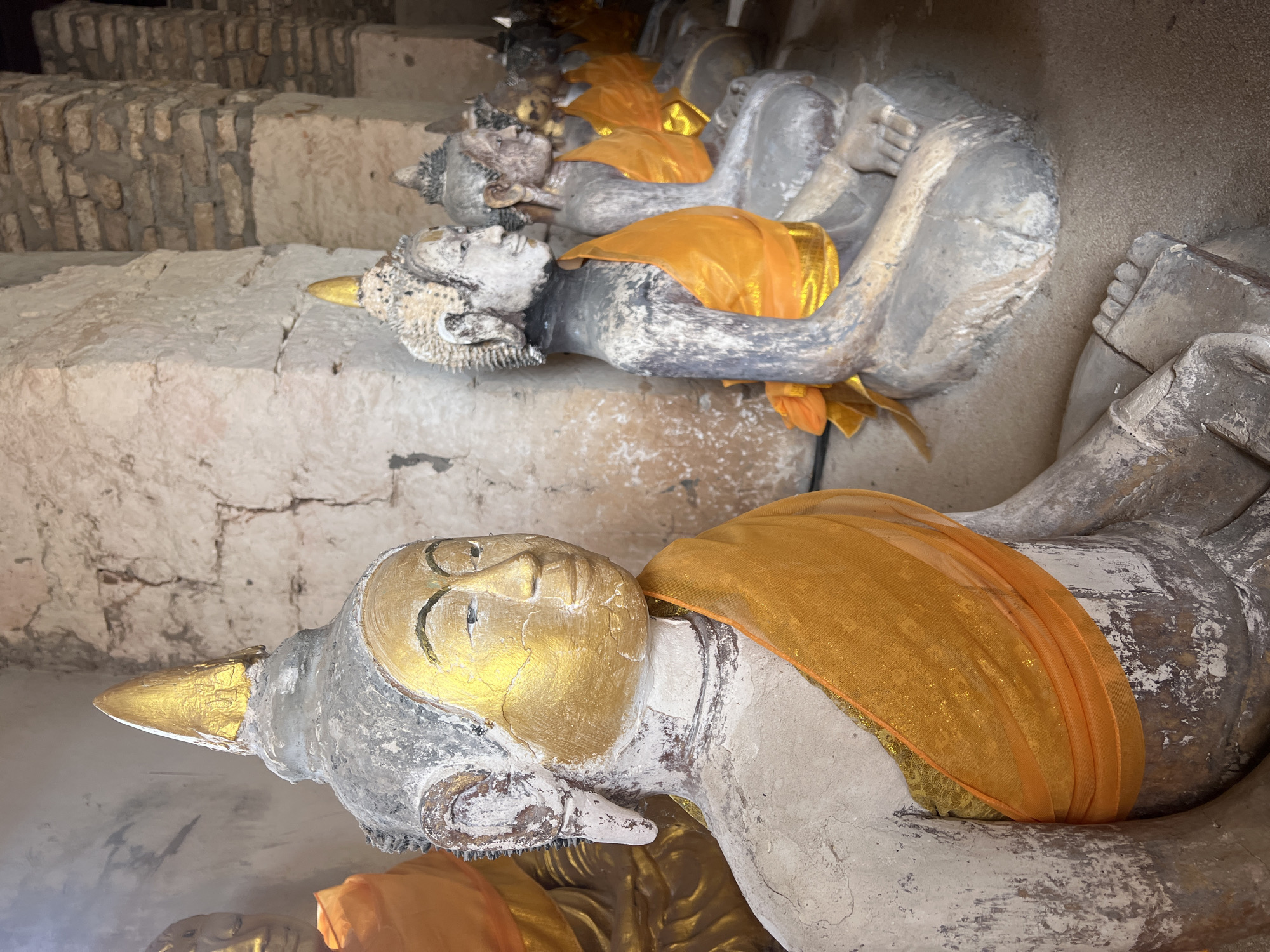 A row of reclining Buddha statues is located at Wat Yai Chaimongkol in Baan Harn, Thailand. The statues are made of stone, are painted white with golden details on the head and adornments. Each statue is draped with an orange fabric. The statues are situated on a series of stone tiers. The stonework exhibits wear and age.