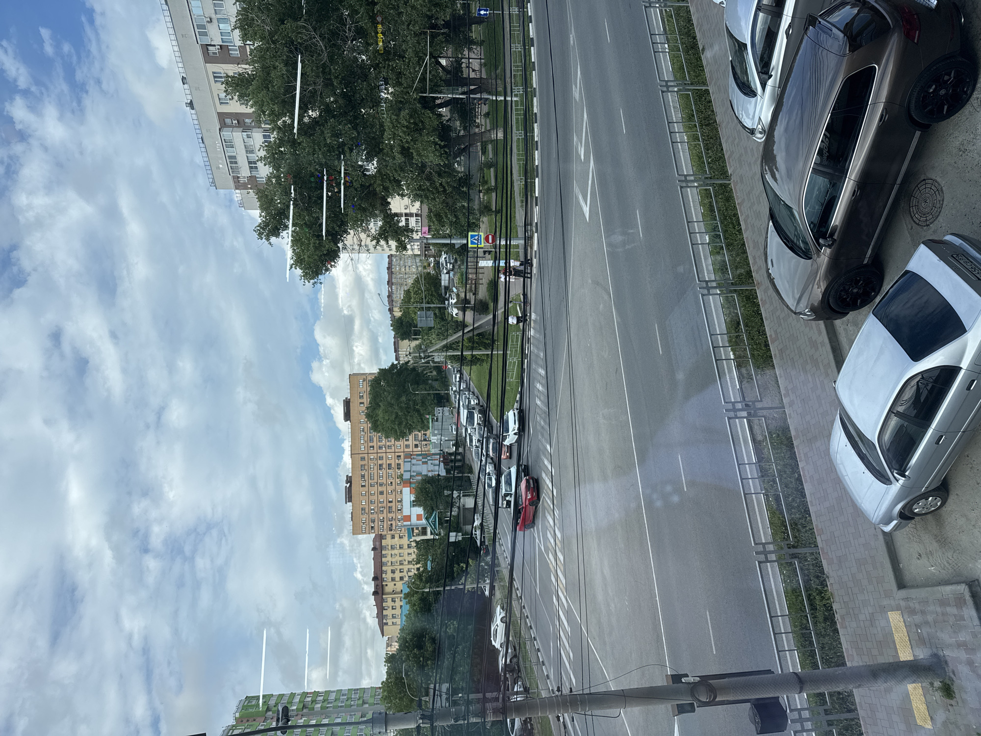 The image shows a view from a window looking out at a city street in Anapa, Russia. A busy intersection with multiple lanes of traffic is visible, with cars moving in various directions. Pedestrian crossings are marked on the asphalt. Several multi-story apartment buildings line the street, with trees and green spaces interspersed. The sky is cloudy. In the foreground, a silver sedan and a dark brown SUV are parked on a paved area next to the sidewalk. A lamppost with a trash receptacle is also in the foreground. The scene appears to be a typical urban environment during the daytime.