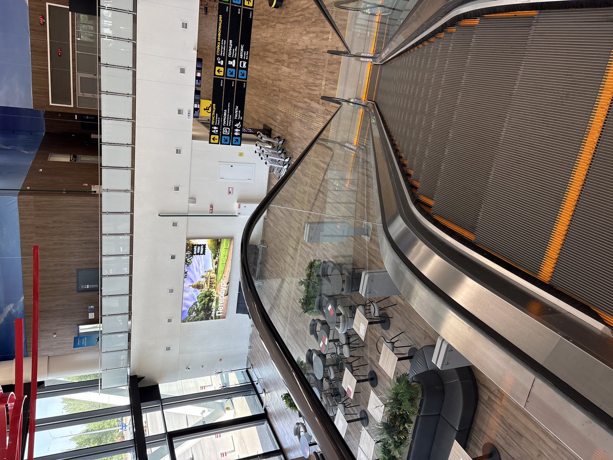 The image shows the interior of an airport, possibly in the city of Новое Мартьяново, Russia. There is an escalator moving away from the viewer, with a gray surface and orange lines. Next to the escalator, there is a seating area with round tables and chairs, along with some greenery. A curved glass barrier separates the seating area from the escalator. Signage in Russian indicating "Information Desk", "Police", "Bus Stop", "Registration", "Parking" and "Restrooms" are visible. There is a large advertisement for "Петербург" (Saint Petersburg) on the wall. Luggage trolleys are parked next to the sign. There are also windows allowing natural light into the area, with some trees visible outside.