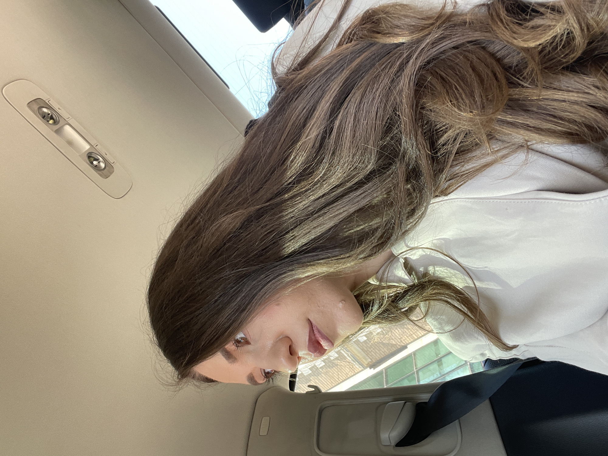 A woman with long brown hair is seated in a car. She is wearing a white top. The interior of the car is light-colored. The car's roof features a light fixture. A seatbelt is visible. The location is unknown.