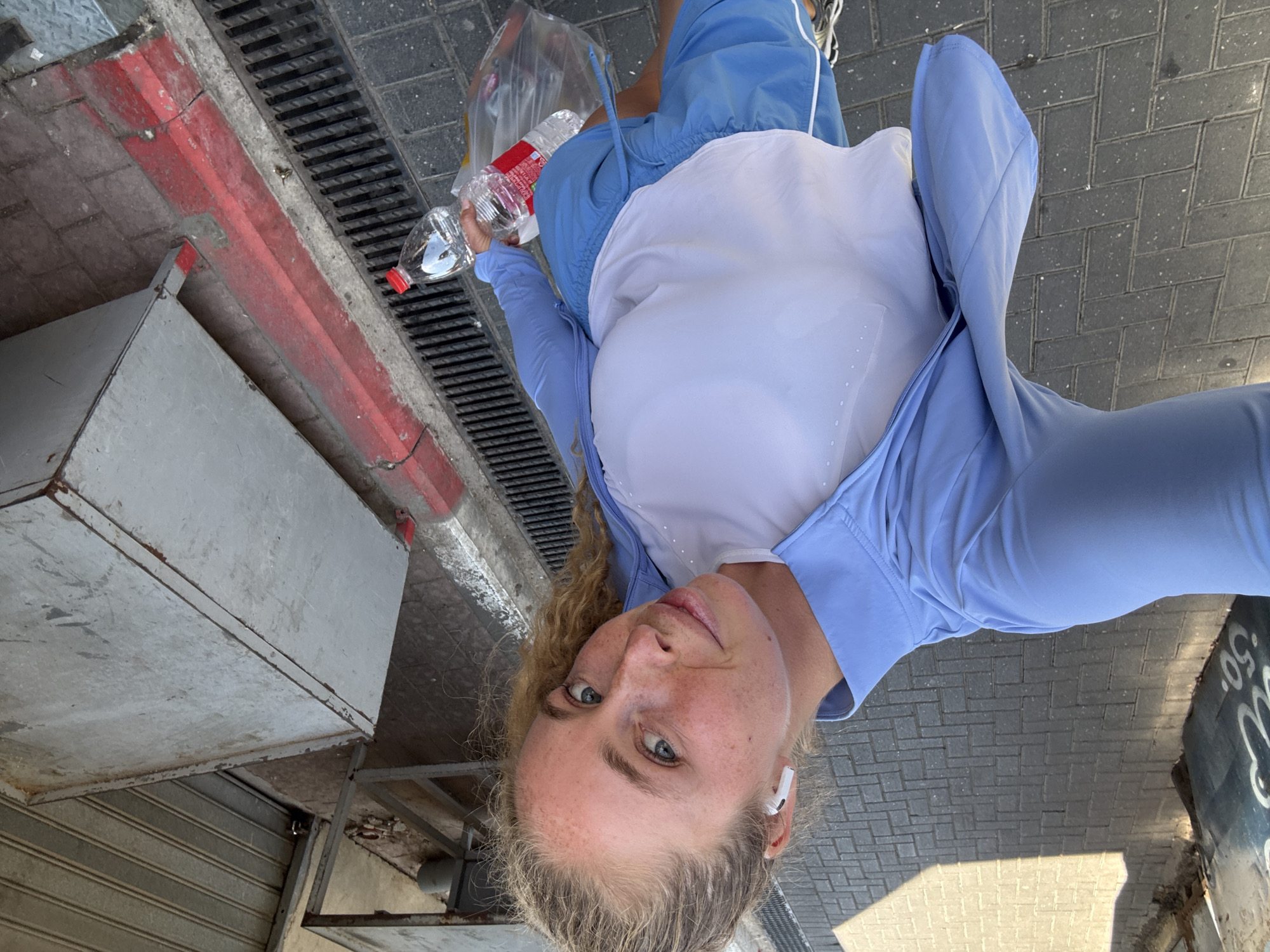 A young woman with blonde hair pulled back in a ponytail and wearing a light blue zippered jacket over a white tank top and blue athletic shorts is taking a selfie outdoors in an urban environment. She has a smattering of freckles across her nose and cheeks and is wearing white wireless earbuds. In her right hand, she holds a plastic bag containing a water bottle. The ground is paved with gray brick-like stones. A red-painted curb runs alongside a metal grate. In the background, there is a corrugated metal shutter and some metallic structures, possibly part of a stall or building facade. The scene appears to be a street in Tel Aviv, Israel, given the context of the location.