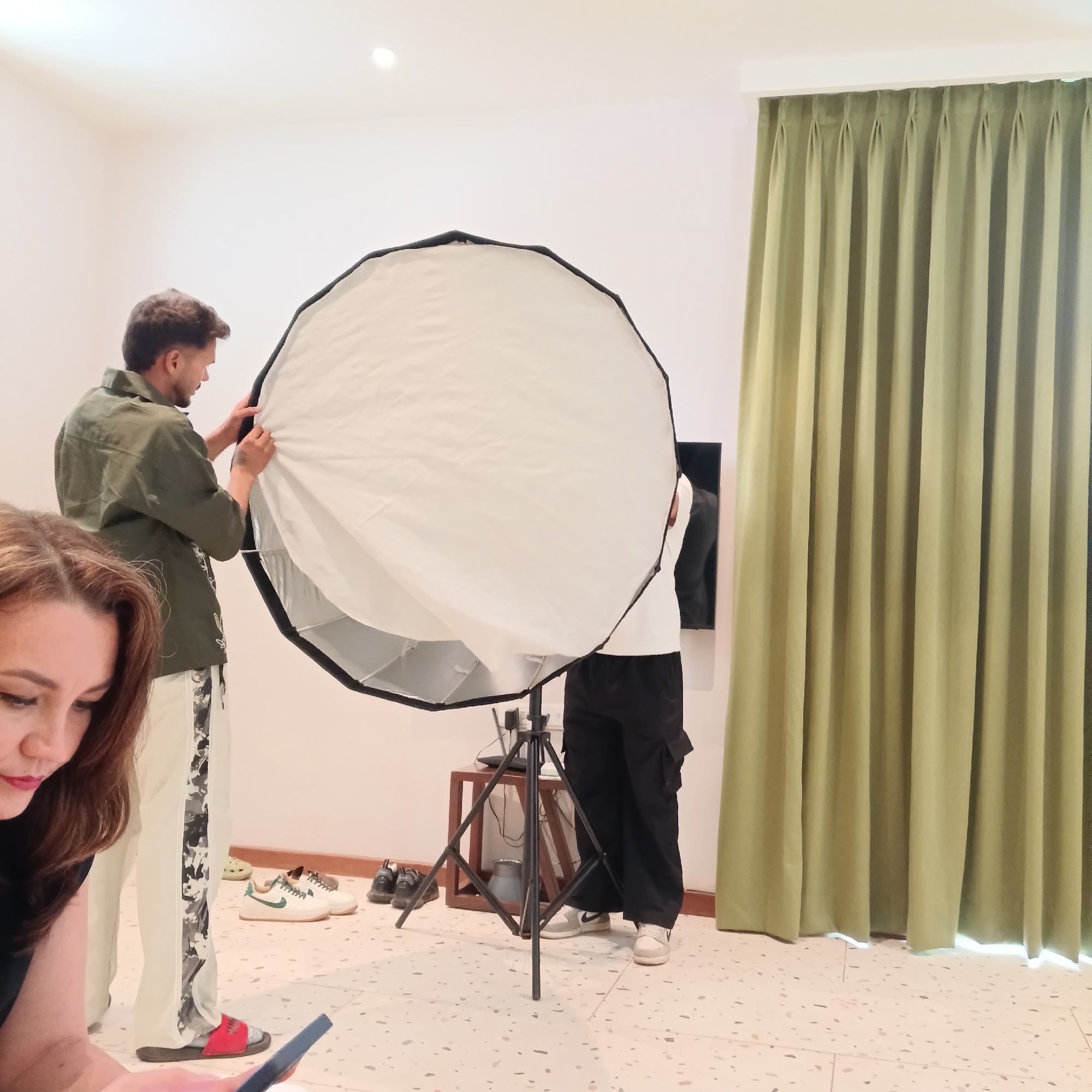 The scene appears to be a studio or indoor setting, potentially in Calangute, India. Two people are assembling a large, circular softbox on a tripod. One person, wearing an olive green shirt and white pants with a camouflage stripe, is working on the front of the softbox. Another individual, dressed in a white shirt and black cargo pants, is positioning the softbox. A woman is visible in the foreground, looking at a cell phone. Several pairs of shoes are scattered on the floor next to the softbox. A small wooden table is nearby, holding additional equipment. Green curtains are drawn on the right side of the frame, next to a black monitor or TV hanging on the wall. The floor has a speckled pattern.