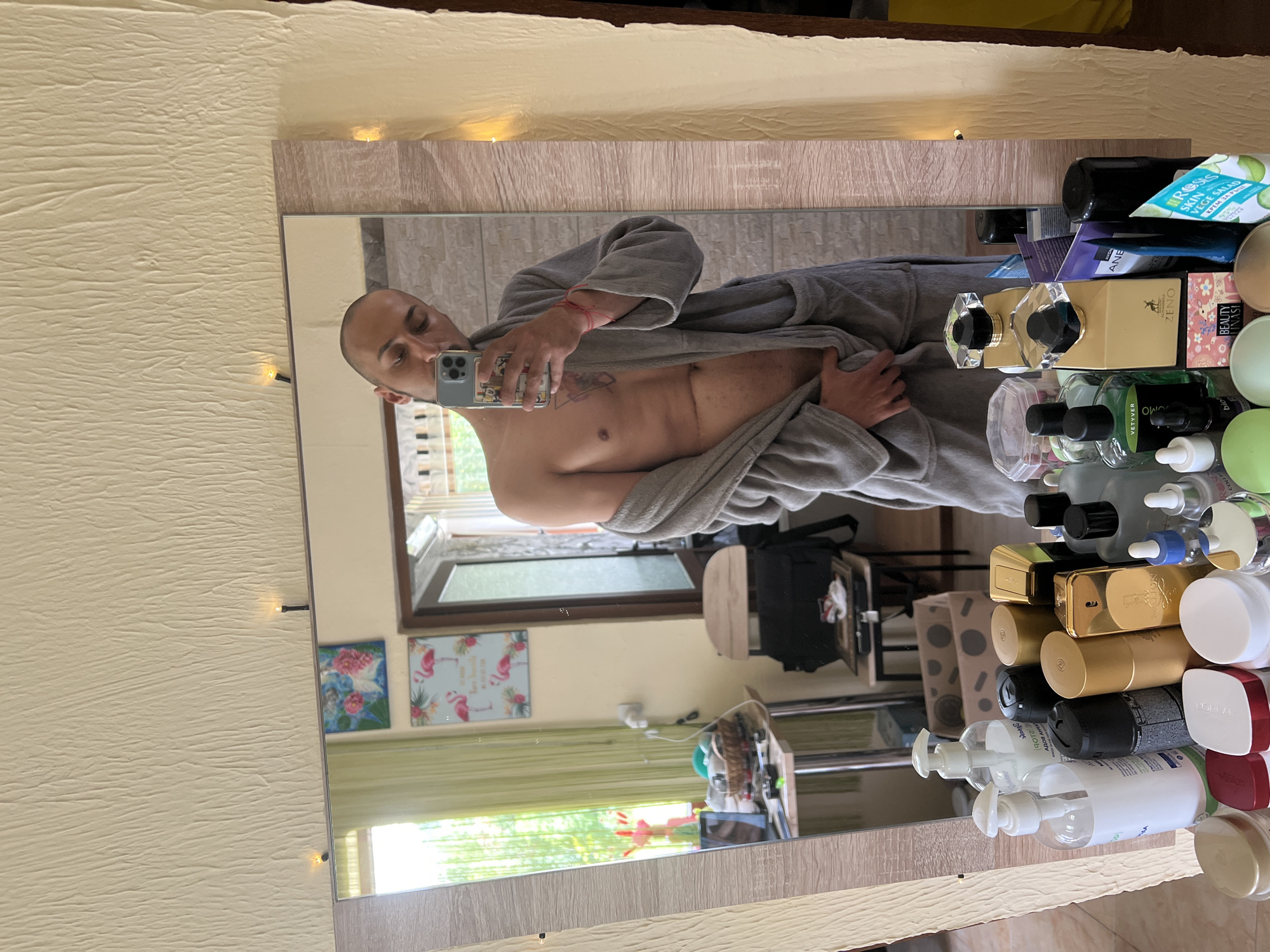 A person is depicted taking a mirror selfie in what appears to be a bathroom or dressing area. The individual, a man with a shaved head, is looking towards their smartphone held up to capture their reflection. He is wearing a gray bathrobe, partially open to reveal his bare chest. A scar is visible on his right chest, and a tattoo is present on his left rib cage. He also wears a red string bracelet on his right wrist. His phone case has a cartoon character design.
In front of the mirror, numerous cosmetic and perfume bottles are arranged on a surface. Visible text on these products includes "VETYVER", "ANE", "ROSES", "SKIN", "VEGE SALAD", "КРЕМ ЗА РЪЦЕ" (Hand Cream), "ZENO", "bio", "BEAUTY INASI", "COLL", "YOPLA", "АДОВ АНЧА", and a partial "E2106". A red-capped container appears to have "L'OREAL" branding.
The mirror's reflection shows a room with light, textured walls. A window in the background reveals outdoor greenery and daylight, indicating it is daytime. Two framed pictures, one with pink flamingos and another with flowers, are visible on the wall behind the person. Other reflections include furniture and bags, contributing to the sense of a personal indoor setting. The scene offers a casual glimpse into a personal space, located in Medkovets, Bulgaria.