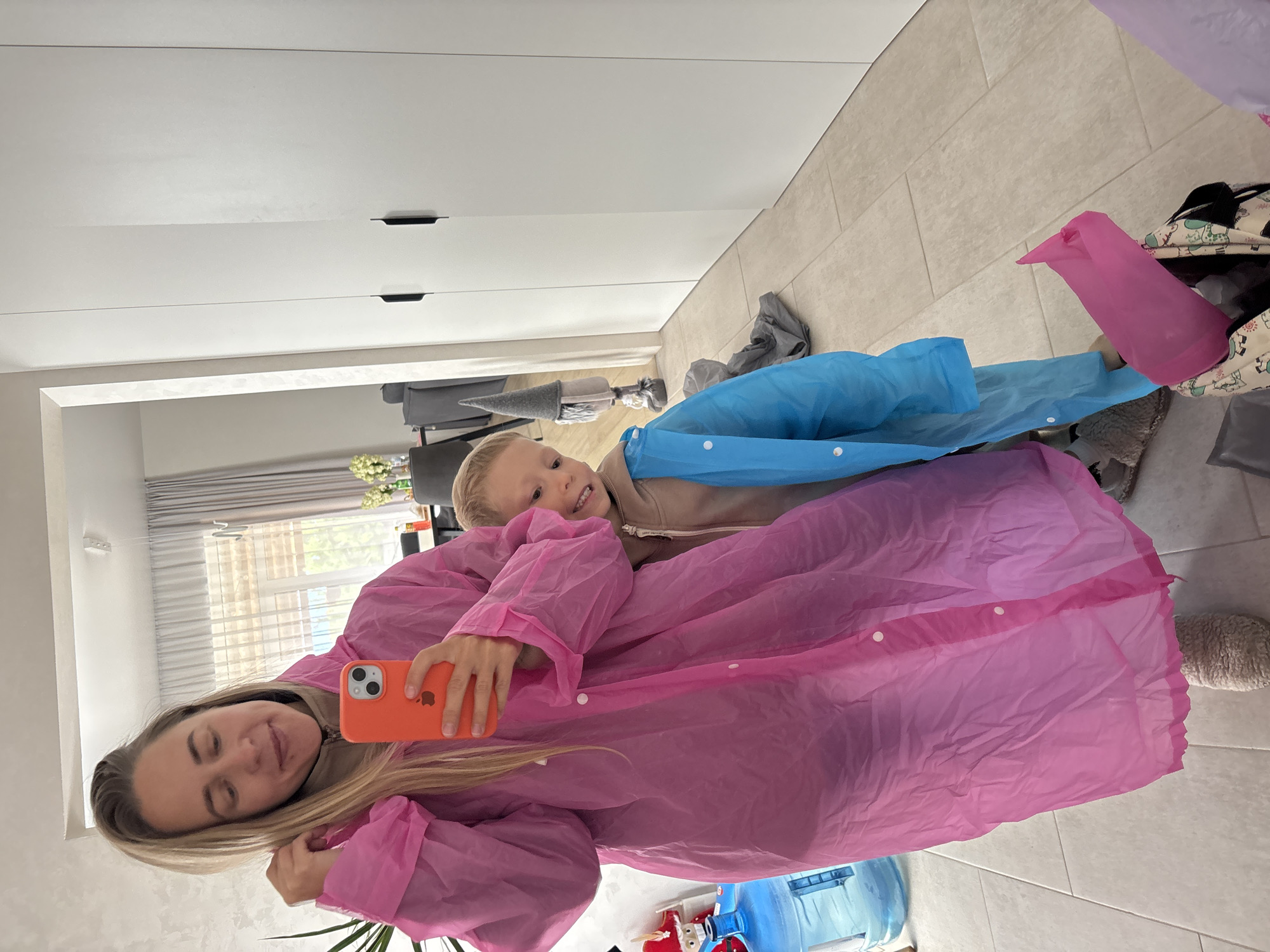 A woman and a young boy are taking a selfie in what appears to be a residential hallway. The woman, with blonde hair pulled back, is wearing a bright pink, oversized, disposable-looking raincoat and holding up an orange smartphone to take the photo. She has a faint smile and is adjusting her hair. The boy, to her right and slightly behind her, is wearing a light blue, similar disposable raincoat and a beige hoodie. He is smiling widely, with his teeth showing, and peeking out from behind the woman. In the background, to the left, there is a large blue water jug. Further left, there is a hint of a potted plant and a window with sheer curtains. To the right of the woman and boy, there is a tall, white, modern-looking wardrobe with dark handles. A decorative gnome figure with a grey pointed hat is visible between the woman and the wardrobe. The floor is tiled with light-colored, rectangular tiles. A pink plastic bag and other items are on the floor in the foreground. The scene is indoors, likely within a home or apartment building, given the wardrobe and hallway-like setting. There is no indication of the specific location within the city of Chaikovsky, Russia.