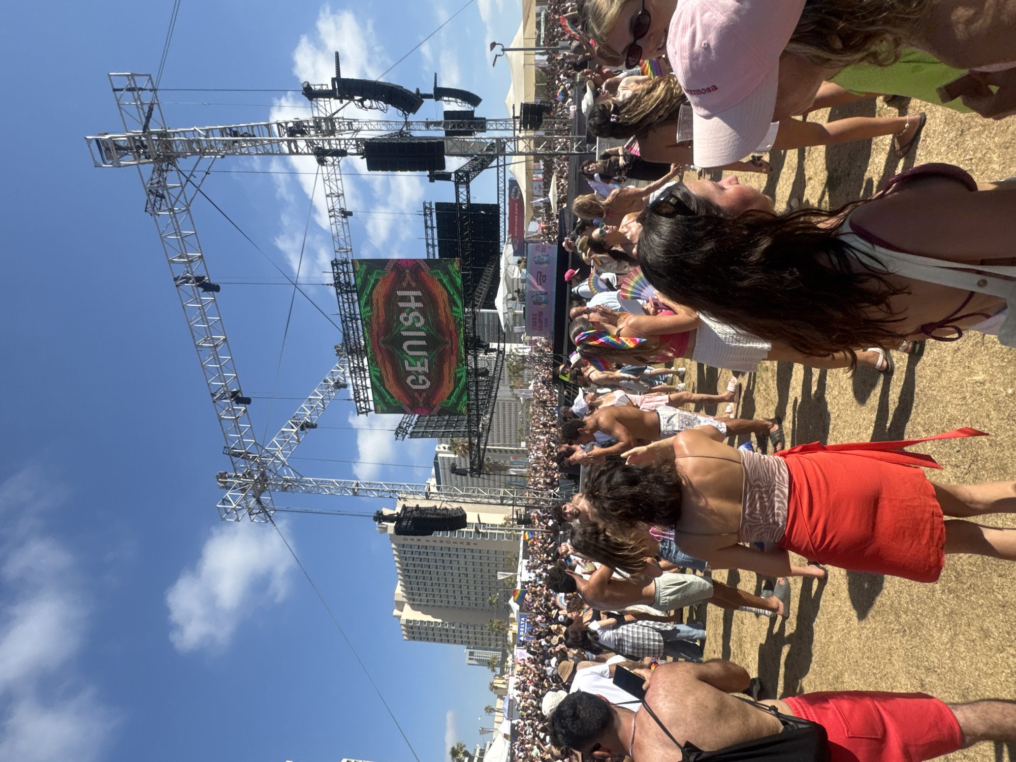 This is a wide shot of an outdoor festival or concert in Tel Aviv, Israel, on a sunny day. A large crowd of people are gathered, facing a stage setup with a large LED screen displaying the word "GENISH" in an artistic font. The screen is framed by a metal truss structure holding speakers and lighting equipment. In the background, tall buildings are visible, characteristic of the Tel Aviv skyline.
The crowd is diverse, with people of various ages and genders. Many are dressed in casual summer attire, and some are holding rainbow-colored items, suggesting this might be a Pride event. People are standing, dancing, and interacting with each other. The ground appears to be covered in dry grass or sand. Tents and smaller structures are set up around the perimeter of the event space. The sky is blue with scattered white clouds.
