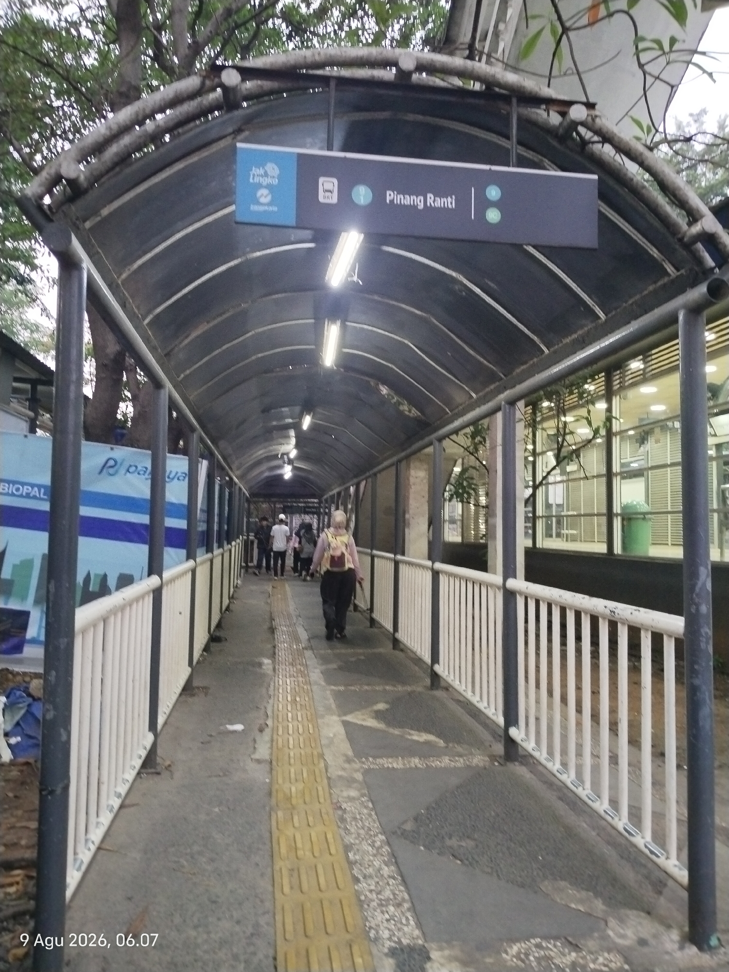 The image shows a covered pedestrian walkway in East Jakarta, Indonesia, near the Pinang Ranti bus station. The walkway is covered by a translucent, curved roof supported by metal poles. A sign indicates "Pinang Ranti" and "BRT" (Bus Rapid Transit). On the left side of the walkway, there is a banner with the text "BIOPAL" and "R paya." The walkway has a tactile paving strip of yellow tiles along the right side. Several people are walking along the path, with one woman in the foreground wearing a backpack and dark pants. Trees are visible above the walkway, and to the right, there are large windows showing an indoor area with fluorescent lighting and a green trash can. The overall scene appears to be an outdoor public transportation area.