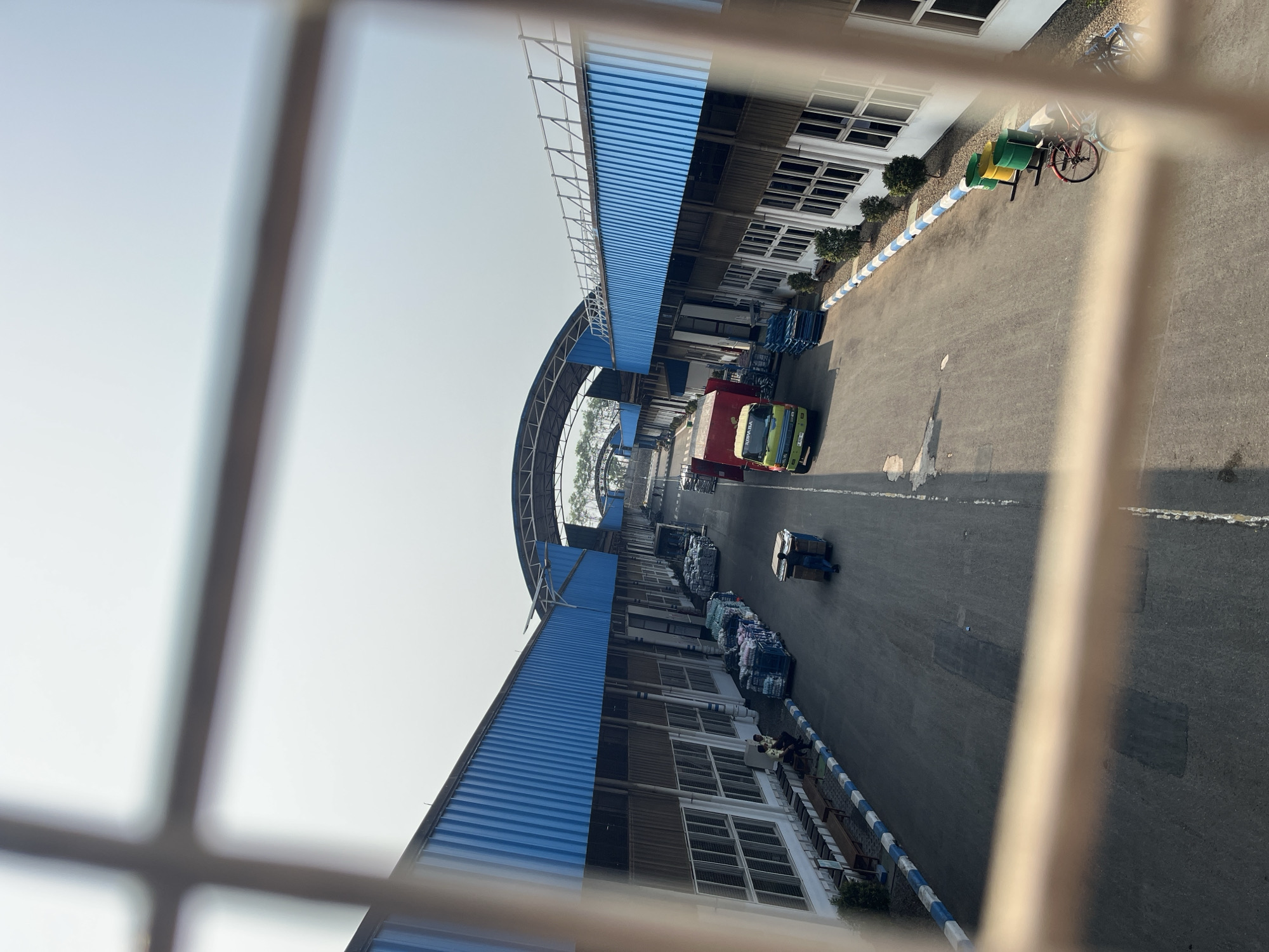 The image shows an outdoor industrial area with buildings on either side of a paved road. The buildings have blue corrugated metal roofs and white walls with numerous windows. The road is lined with blue and white curbs and has several trucks, carts, and stacks of goods. There are also some people visible in the scene. In the foreground, a blurry fence or grate obscures the view. The sky is bright and overcast, suggesting a daytime setting. Based on the provided information, this scene is located in the city of Pelang, Indonesia.