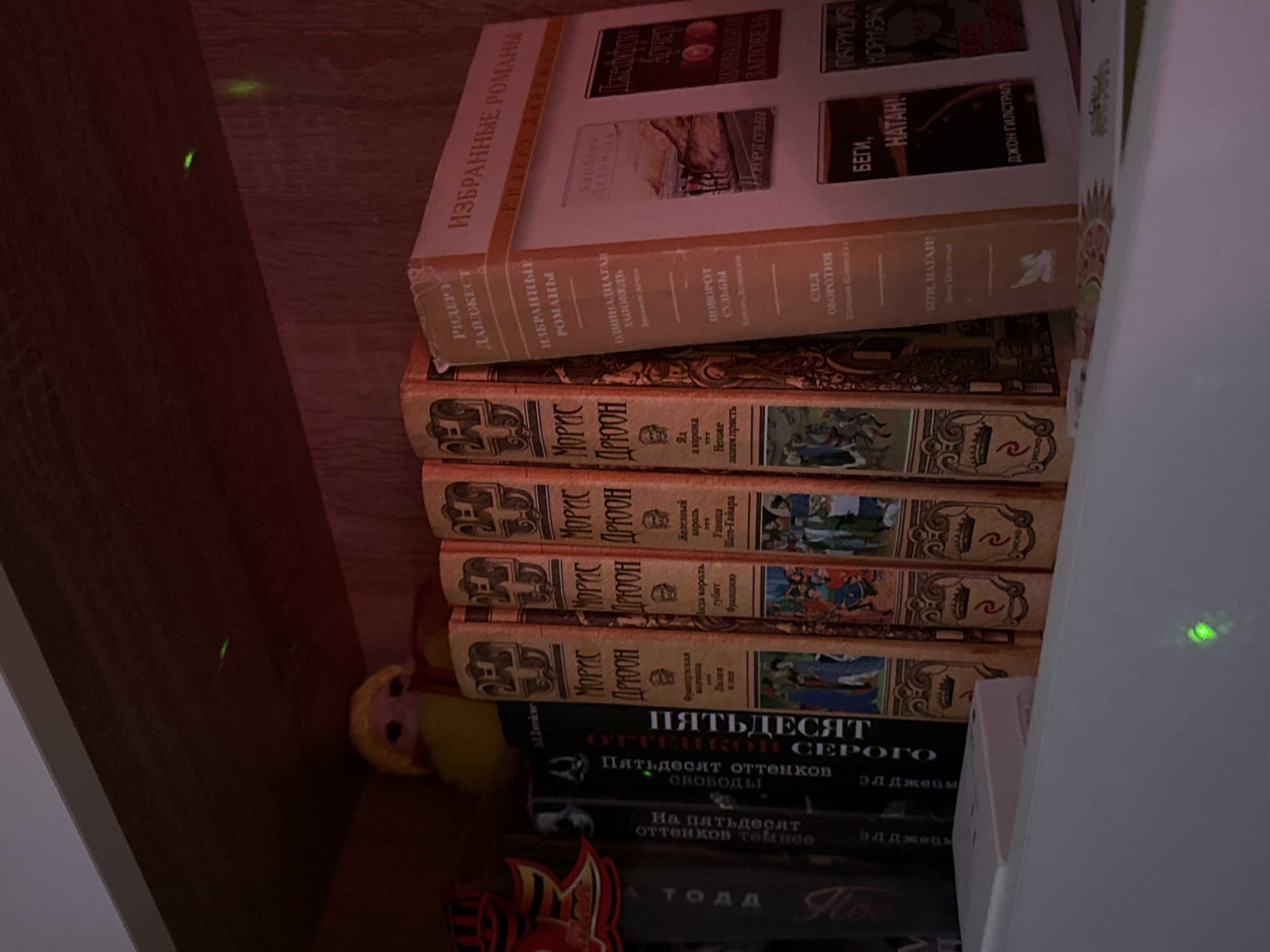 A close-up shot depicts a section of a bookshelf, crammed with books stacked vertically. The top book is a Reader's Digest collection titled "Selected Novels," with a montage of book covers printed on the spine, including titles like "The Eleventh Commandment" and authors like Barbara Delinsky. Beneath this, a series of books by Maurice Druon are visible, distinguished by their dark brown spines adorned with regal crests. Titles such as "The Iron King" and "Lily and Lion" can be deciphered. Further down, "Fifty Shades of Grey" and its sequels are present, their spines dark with white lettering. In the corner of the frame, a small, yellow, plush doll is partially visible. The surrounding shelves are made of wood, and the lighting is dim, casting shadows on the scene.