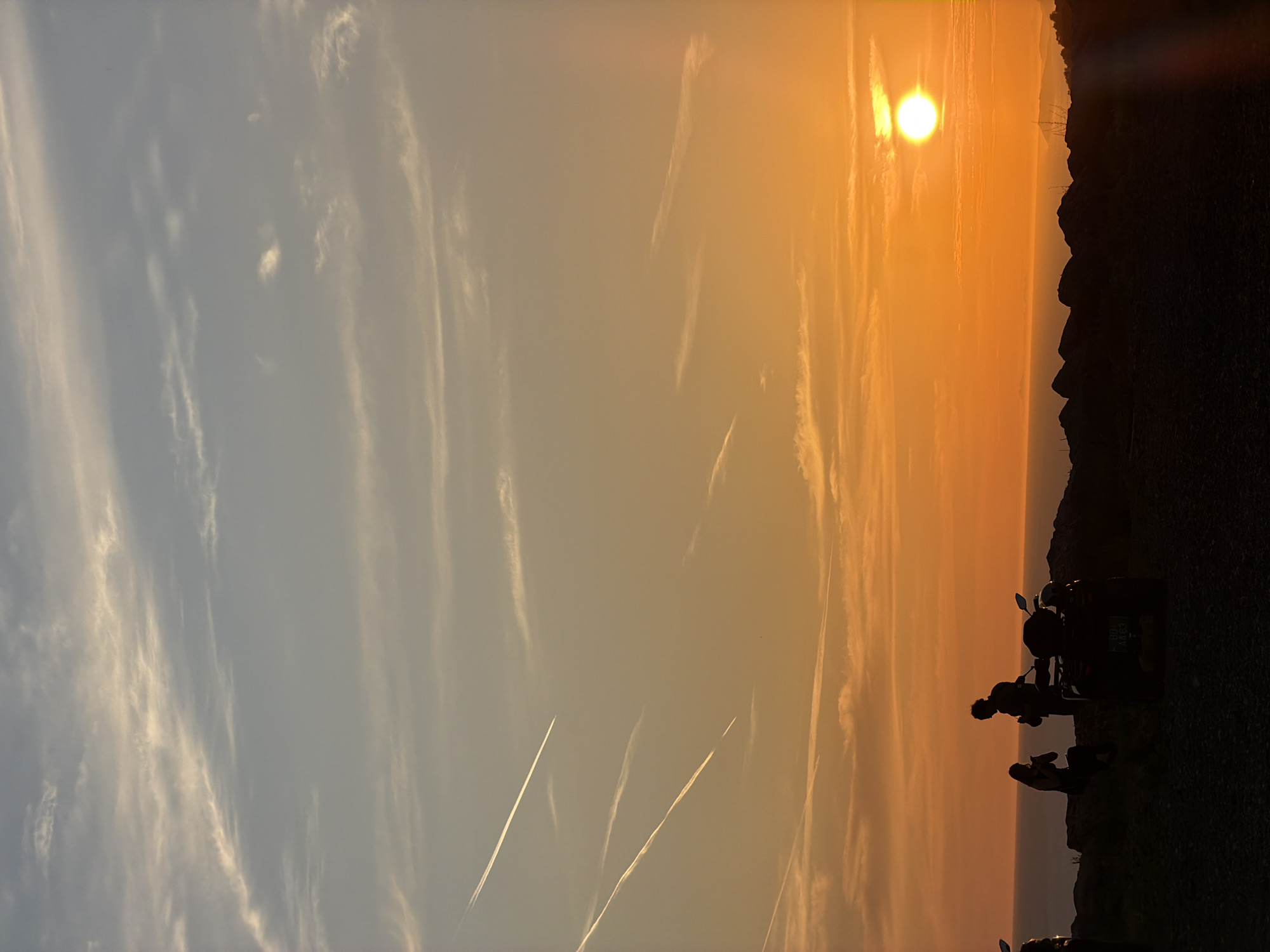The image depicts a sunset scene in Kovačko Polje, Montenegro. In the foreground, silhouetted against the vibrant orange sky, are two people and a quad bike. One person is seated on a rocky outcrop, while the other stands beside them. The quad bike is parked to the right of the figures. The background features the sea meeting a distant, hazy coastline. The sky is adorned with wispy clouds and several distinct contrails from aircraft, streaking across the gradient of orange and blue hues. The overall impression is one of a peaceful, scenic evening in a coastal or elevated landscape.