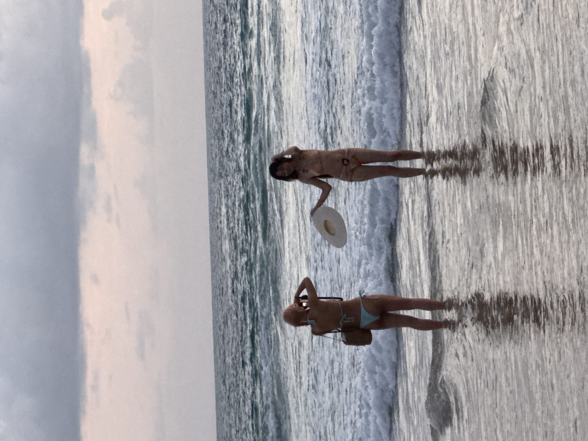 Two women are standing in the shallow water of Karon Beach, Thailand. The woman on the left is wearing a light blue bikini and has a straw bag slung over her shoulder. She is holding a camera and appears to be taking a picture. The woman on the right is wearing an orange bikini and is holding a wide-brimmed straw hat. The water is choppy with small waves and the sky is overcast, suggesting it is either dawn or dusk. The overall scene is natural and candid, capturing a moment of leisure on the beach.