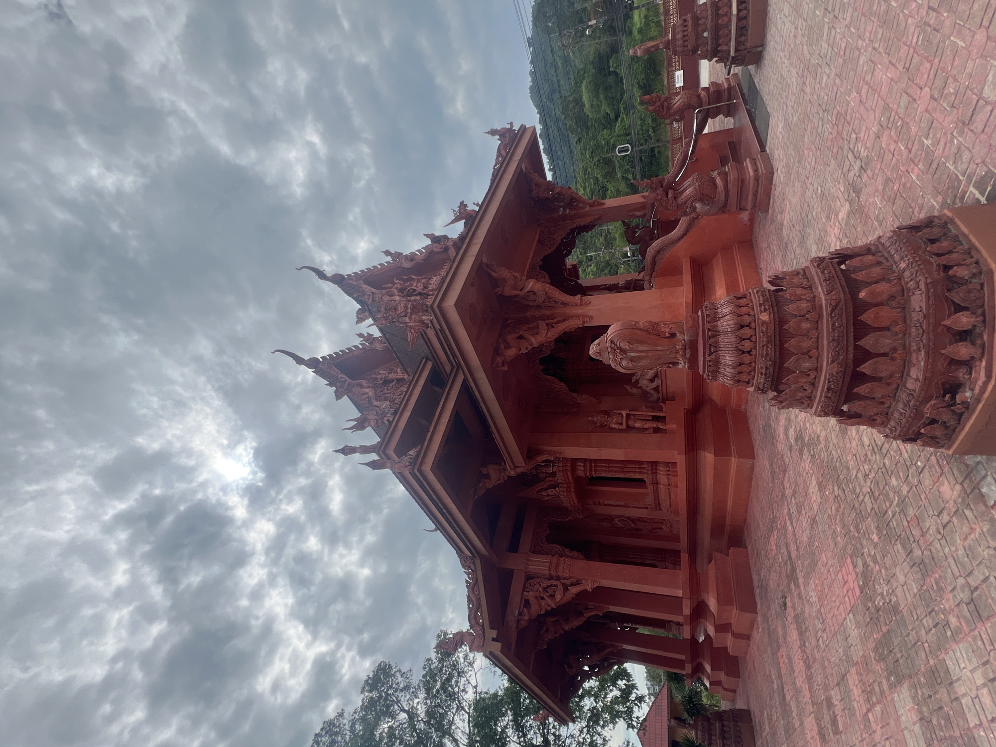 The image shows an ornate, red wooden temple structure against a cloudy sky. The temple is intricately carved with mythological figures and decorative patterns. A tiled walkway, also in shades of red, leads up to the entrance of the temple. Lush green trees and a hillside can be seen in the background, suggesting a natural or mountainous setting. The sun is partially visible through the clouds, indicating it is daytime. There are no people visible in the frame, and no text is discernible. The overall atmosphere is serene and spiritual, emphasizing the detailed architecture of the temple.