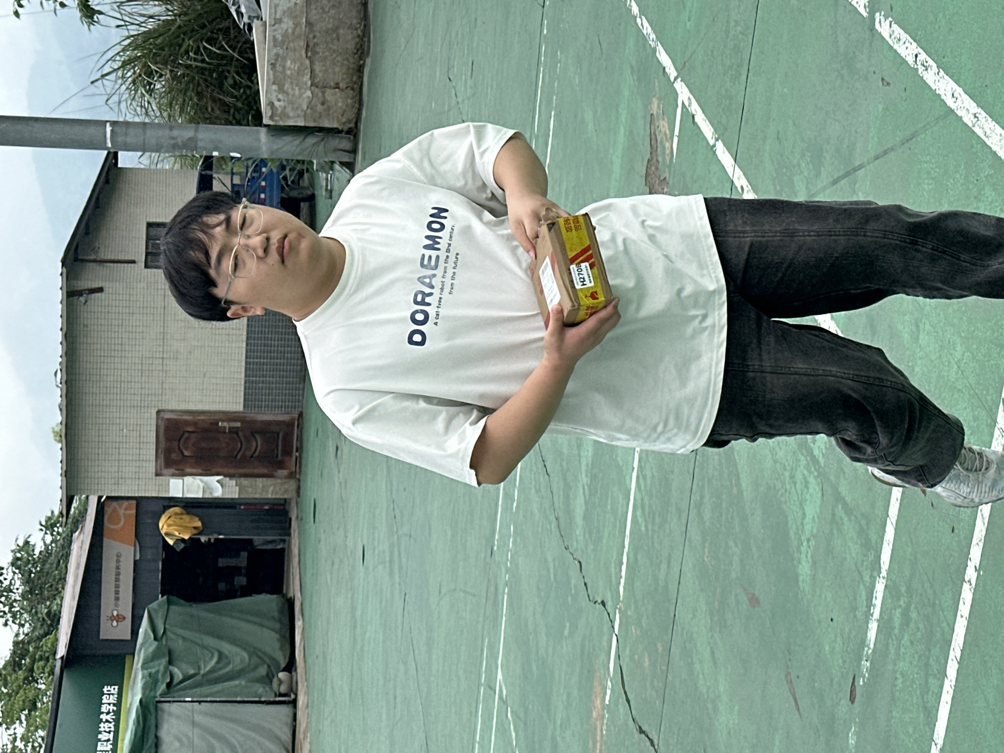 A person wearing glasses, a white Doraemon t-shirt, and dark-colored jeans is holding a cardboard box in their hands on a green surface marked with white lines, possibly a sports court. The box has text, including the code "H2708". Behind the person is a building with a dark doorway partially covered by a green sheet or tarp. A sign above the doorway includes Chinese characters, indicating "星职业技术学院店", possibly meaning a vocational school or shop. The environment suggests an outdoor location in Jiangjin, China.