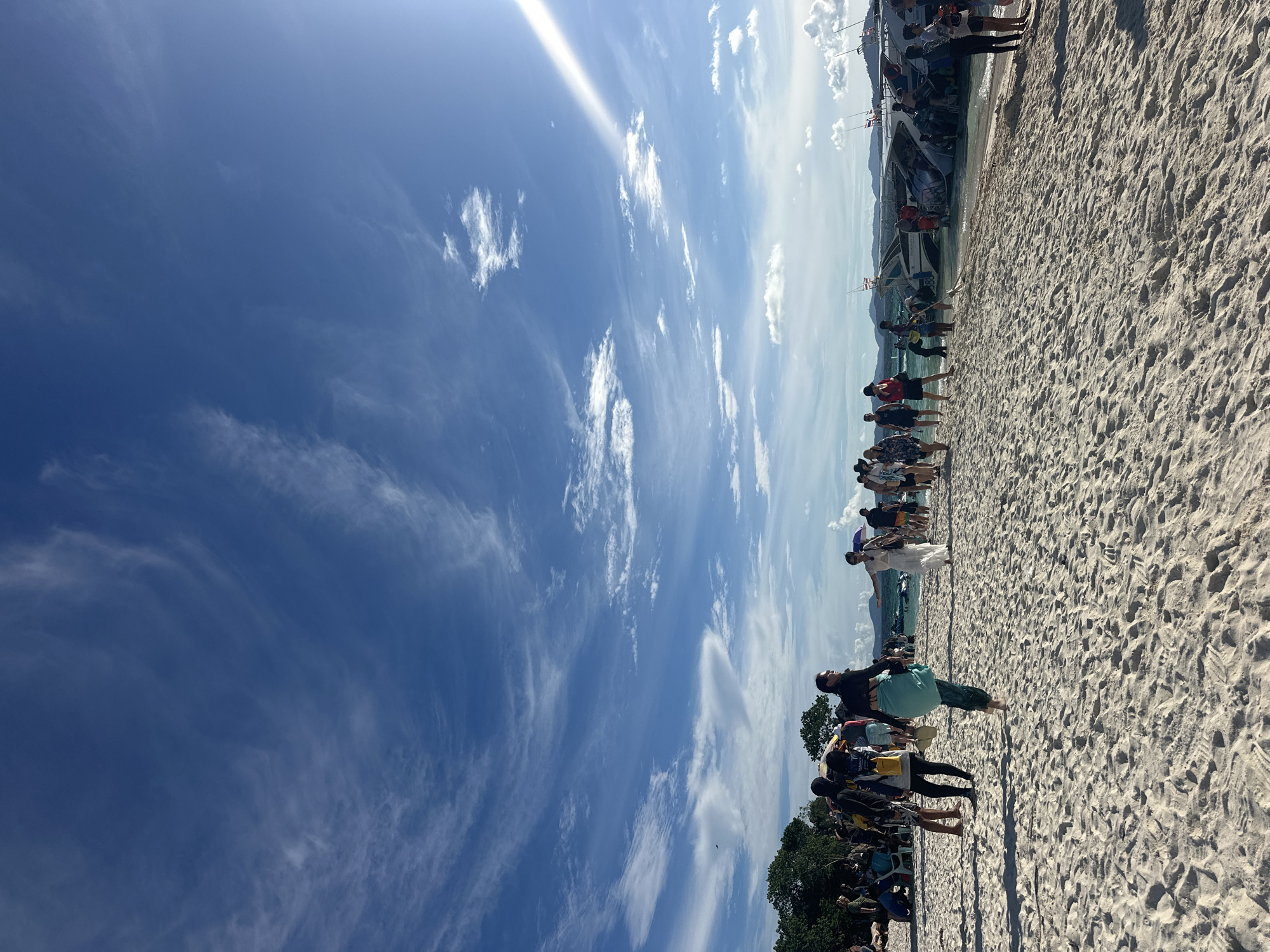 The image depicts a vibrant scene on a sandy beach, likely in Phru Nai, Thailand. The sky is a brilliant blue, scattered with wispy white clouds, suggesting a clear and sunny day, possibly mid-morning or early afternoon given the bright sunlight.
A group of people are gathered along the shoreline, with a speed boat moored nearby. Some individuals are standing, while others appear to be in the process of boarding or disembarking from the boat. Many are dressed in casual, beach-appropriate attire. The mood seems relaxed and perhaps excited, typical of a tourist excursion or day trip.
The beach itself is characterized by fine, light-colored sand, with subtle footprints indicating recent activity. In the background, lush green trees and distant hills can be seen, hinting at a tropical island setting. The water appears calm and shallow near the shore, with the speed boat indicating a mode of transport for exploring the area. Small flags, possibly national flags, are visible on the boat. There is no visible text in the image.