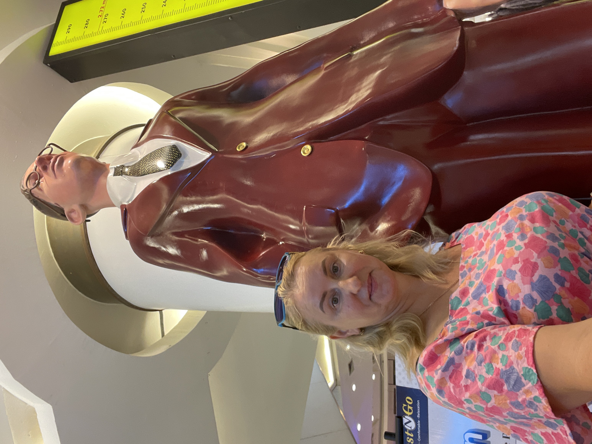 A fair-skinned woman with blonde hair and sunglasses perched on her head is standing next to a very tall, life-sized statue of a man wearing a dark red suit, white shirt, and patterned tie. The statue is so large that it towers over the woman. The statue is glossy and appears to be made of plastic or a similar material. To the right of the statue, a yellow illuminated sign with black markings displays what appears to be a measurement scale, with numbers ranging from 240 to 290. In the background, there is a curved ceiling and architectural elements suggesting an indoor public space, possibly a mall. The woman is wearing a colorful, floral-patterned dress. The overall impression is a snapshot taken in a brightly lit indoor environment.