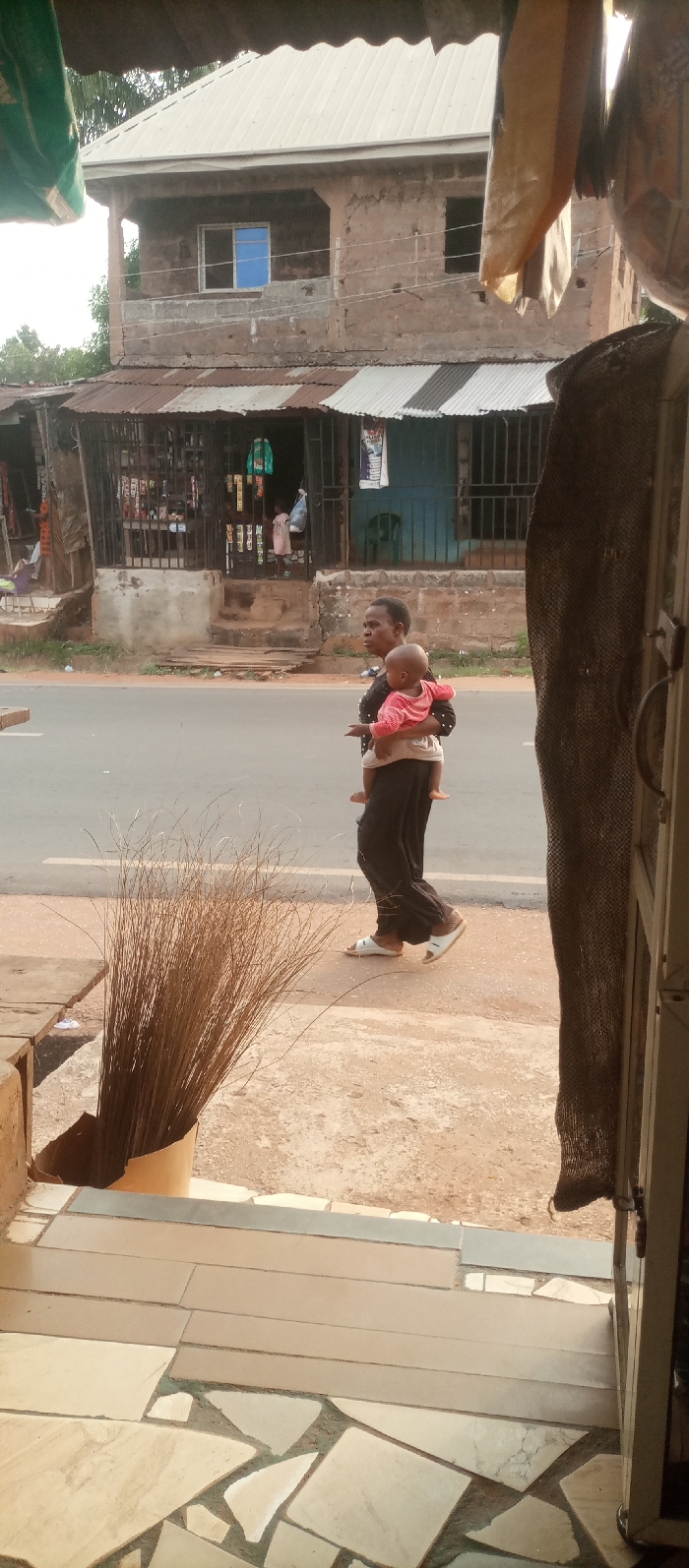 The image portrays a daytime street scene in Ogidi, Nigeria. In the foreground, on a multi-tiled floor, a bundle of dried, broom-like stalks stands upright in a makeshift light-colored container. A partial wooden surface is visible to the left, and a dark mesh fabric hangs from a metallic doorframe on the right.
In the midground, a dark-skinned woman, wearing a dark top with light speckles and dark trousers, walks from left to right on a concrete sidewalk. She is wearing white slip-on sandals. She carries a light-skinned infant, dressed in a pink long-sleeved top, on her right hip. The woman looks forward, slightly to her left.
Behind the sidewalk, an asphalt road with faded white markings runs horizontally across the frame. Beyond the road, a row of single-story commercial stalls with corrugated metal roofs is visible. These shops feature metal grate facades, through which various merchandise, including hanging clothing, can be seen. Two small children, one in a light pink dress, are standing inside the central shop.
In the background, a two-story building constructed of unplastered reddish-brown brick stands behind the stalls. This building features a light-colored corrugated metal roof. The upper floor shows one window with a blue frame and another open window aperture. Green foliage is visible beyond the buildings. Overhead, a section of a light-colored corrugated roof is visible, with beige fabric or sacks hanging down on the right side.