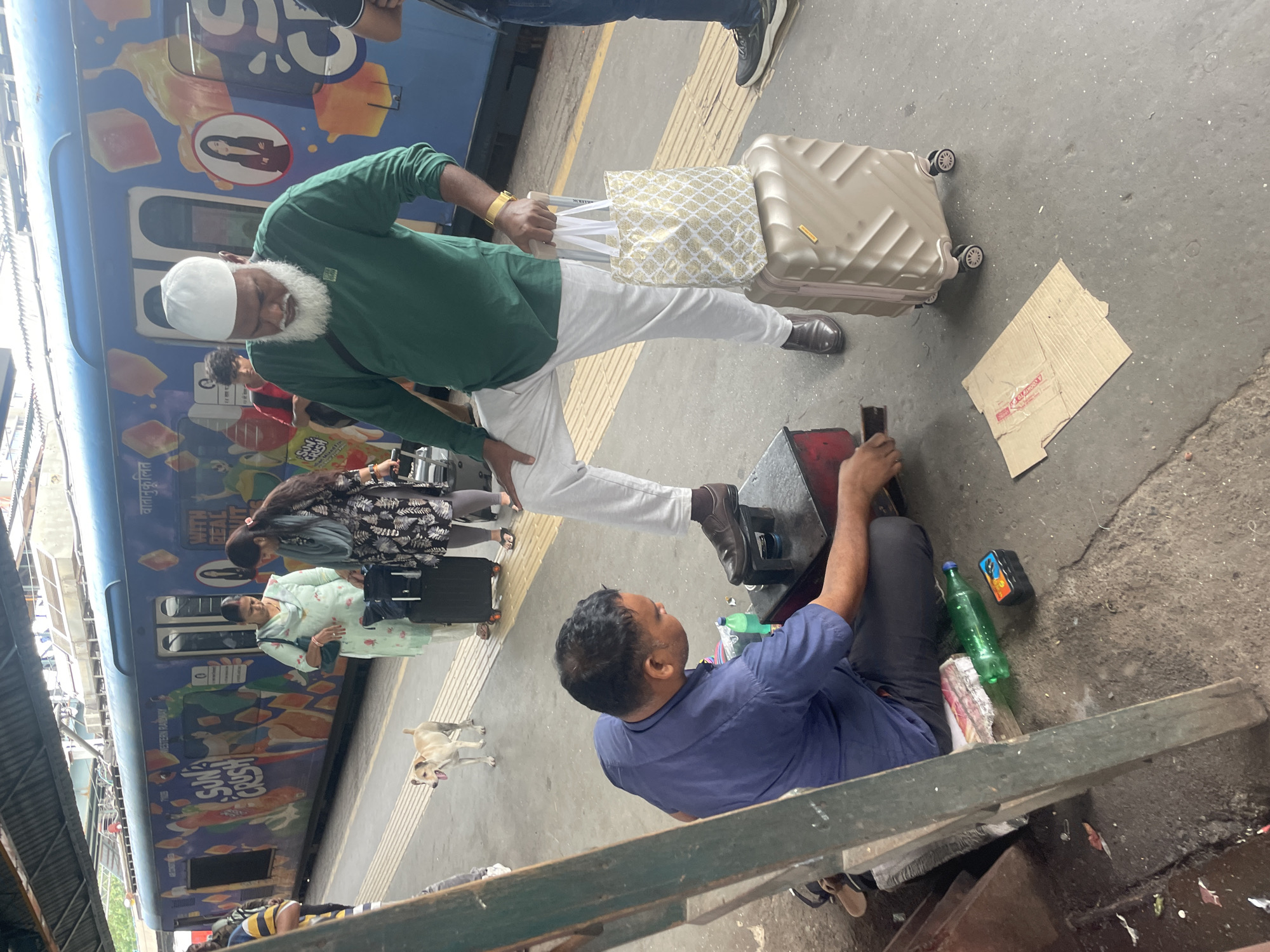This image captures a scene at a busy Mumbai train station. In the foreground, a shoe shiner in a blue shirt is crouched down, meticulously polishing a dark brown shoe that is placed on a small wooden stool. Next to him, there are several items related to his trade, including bottles of polish and a small brush. A man wearing a white skullcap and a green long-sleeved shirt stands with one leg raised, his shoe undergoing the polishing service. He is holding a patterned tote bag with white handles, which rests on top of a beige wheeled suitcase. The background is dominated by a vibrant blue train adorned with colorful advertisements, likely promoting beverages, and featuring the text "SUN CRUSH WITH REAL FRUIT". Several other people are visible on the platform, including women with luggage and a dog, suggesting the general bustle of a public transportation hub. The ground is a concrete platform, typical of Indian train stations, with yellow safety markings along the edge. The overall atmosphere is that of daily life in a bustling Indian city.