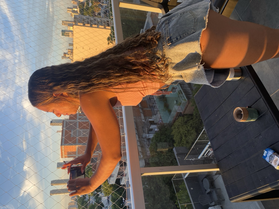 A young woman with long, wavy brown hair stands on a balcony taking a photo with her phone. She wears a pink top and distressed denim shorts. The balcony offers a cityscape view bathed in golden sunlight. A black table holds a metal can, duct tape, and a small package. A protective net covers the balcony railing.