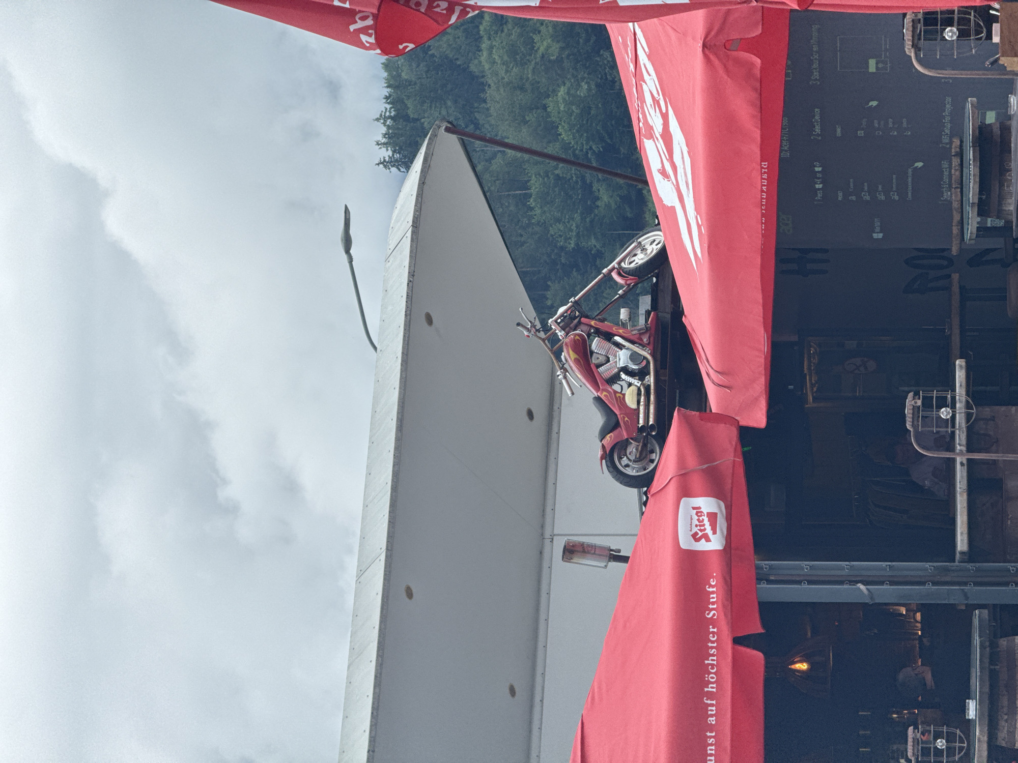 This is a medium shot, outdoor, eye-level photograph taken on a cloudy day in Innsbruck, Austria. A red motorcycle with flame decals is parked on a platform, partially covered by a red umbrella with a white logo. The umbrella displays the text "Stiegl" and German text "Kunst auf höchster Stufe." In the background, a white structure with a curved roof extends across the frame. Trees are visible behind the structure. Below the umbrella, a dark structure with tables and seating is visible. Two people are seated at tables, and industrial-looking lamps are present. A black structure with text and diagrams is also visible in the lower right quadrant of the image, partially obscured by the red umbrella.