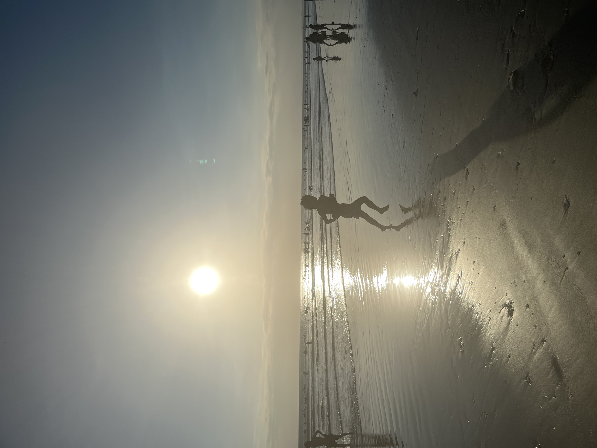 In Kuta, Indonesia, a child is silhouetted against the setting sun as they run along the wet sand of the beach. The sun, a bright orb, is low in the sky, casting a golden glow across the scene and reflecting off the water. In the background, other beachgoers are visible, some wading in the shallow water, while others are further out in the ocean. The waves are gentle, lapping at the shore, and the sand is marked with the footprints of those who have walked there. The overall atmosphere is one of peaceful enjoyment of a beautiful day at the beach.
