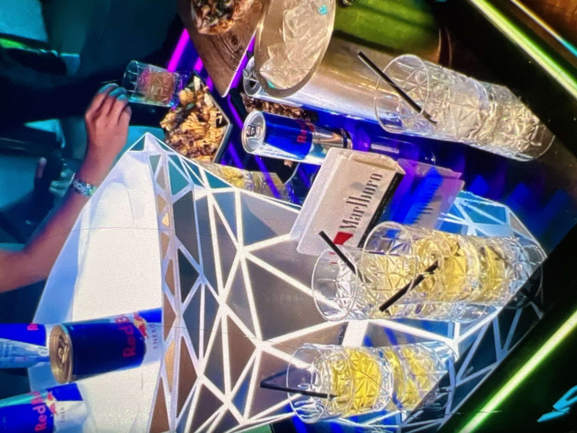 In a dimly lit, vibrant setting, likely a Lagos nightclub or lounge, a low table features a variety of items associated with a night out. Prominently displayed is a white, geometrically patterned ice bucket filled with several cans of Red Bull. Adjacent to it, a tray holds two glasses of amber liquid with straws, suggesting cocktails or spirits. A pack of Marlboro cigarettes rests beside them. Further along the table, a can of Red Bull stands next to a silver ice bucket, which contains ice and a straw. Another tall glass, filled with ice and a straw, sits to the right. In the background, a person's arm, adorned with a bracelet, is reaching towards the drinks, possibly preparing to serve or take one. The overall ambiance is one of leisure and revelry, characteristic of urban nightlife in Lagos.